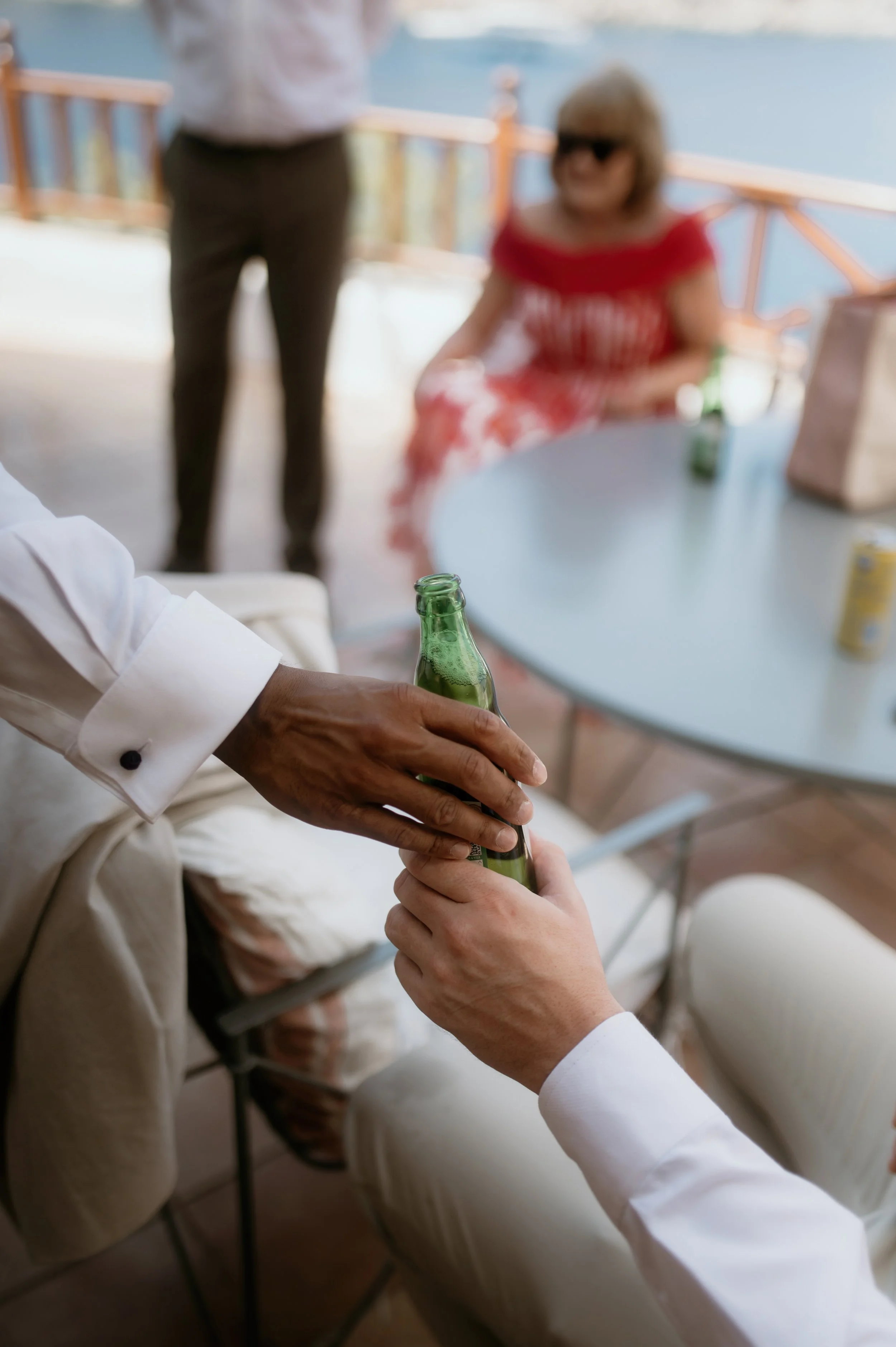Two men holding beer bottles, one sitting and the other standing, with outdoor scenery in the background. A woman in a red dress and sunglasses is sitting at a table behind them.