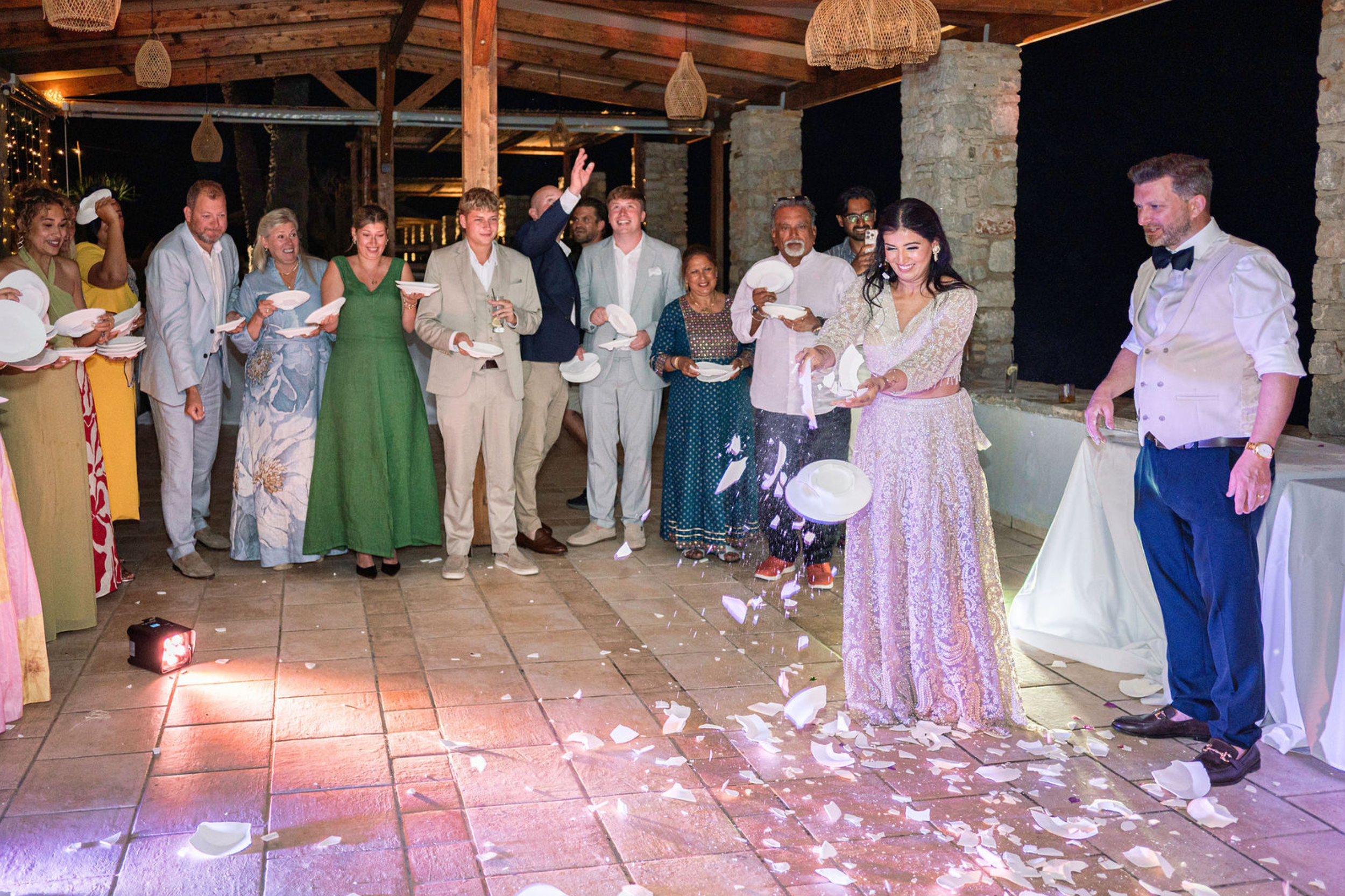Celebration with a group of people at a party, including a woman in a gold dress, a man in a tuxedo, and others standing in the background, with some holding plates and glasses.