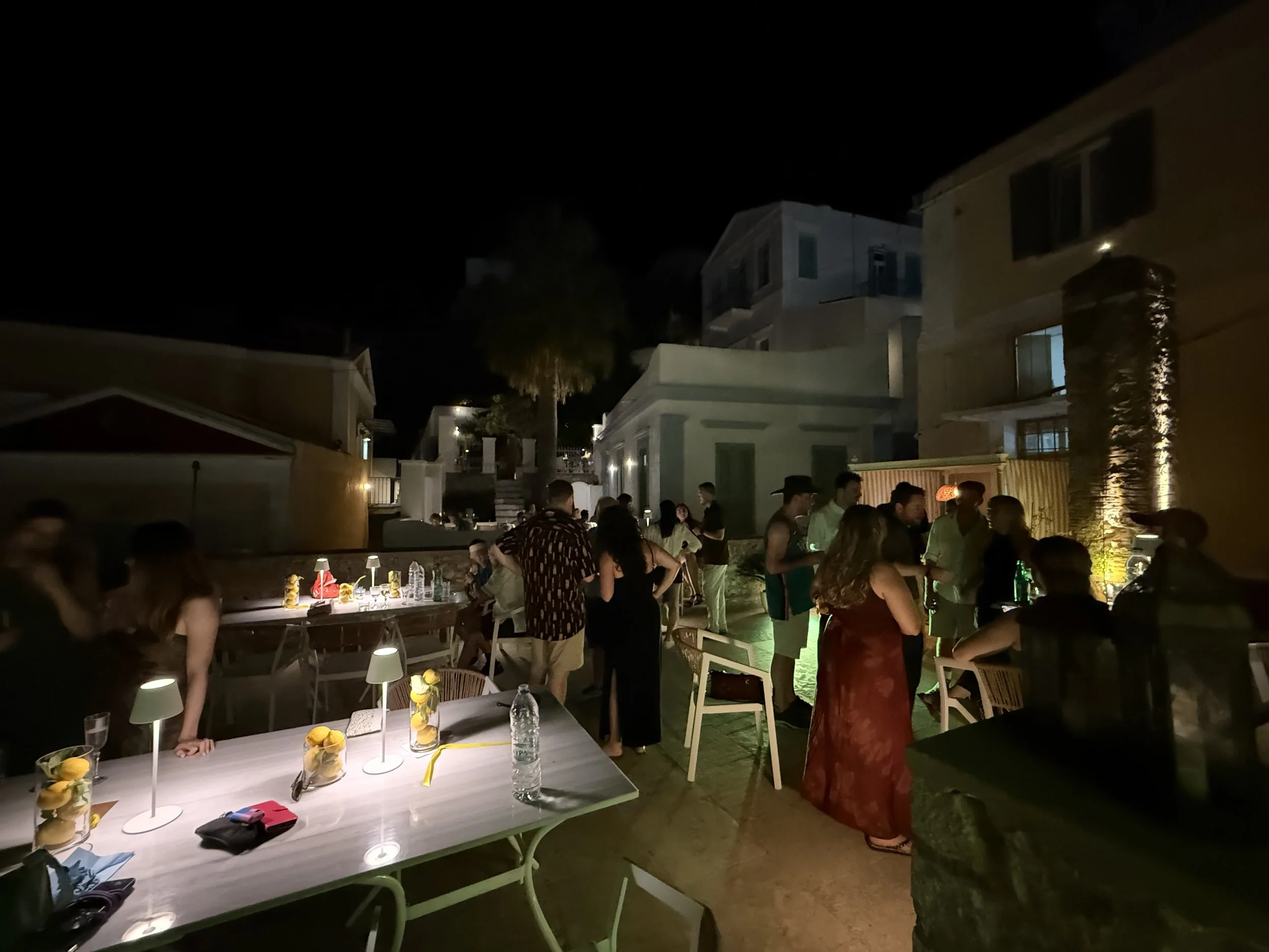 A nighttime gathering with people socializing outdoors, illuminated tables with centerpieces, and houses in the background.