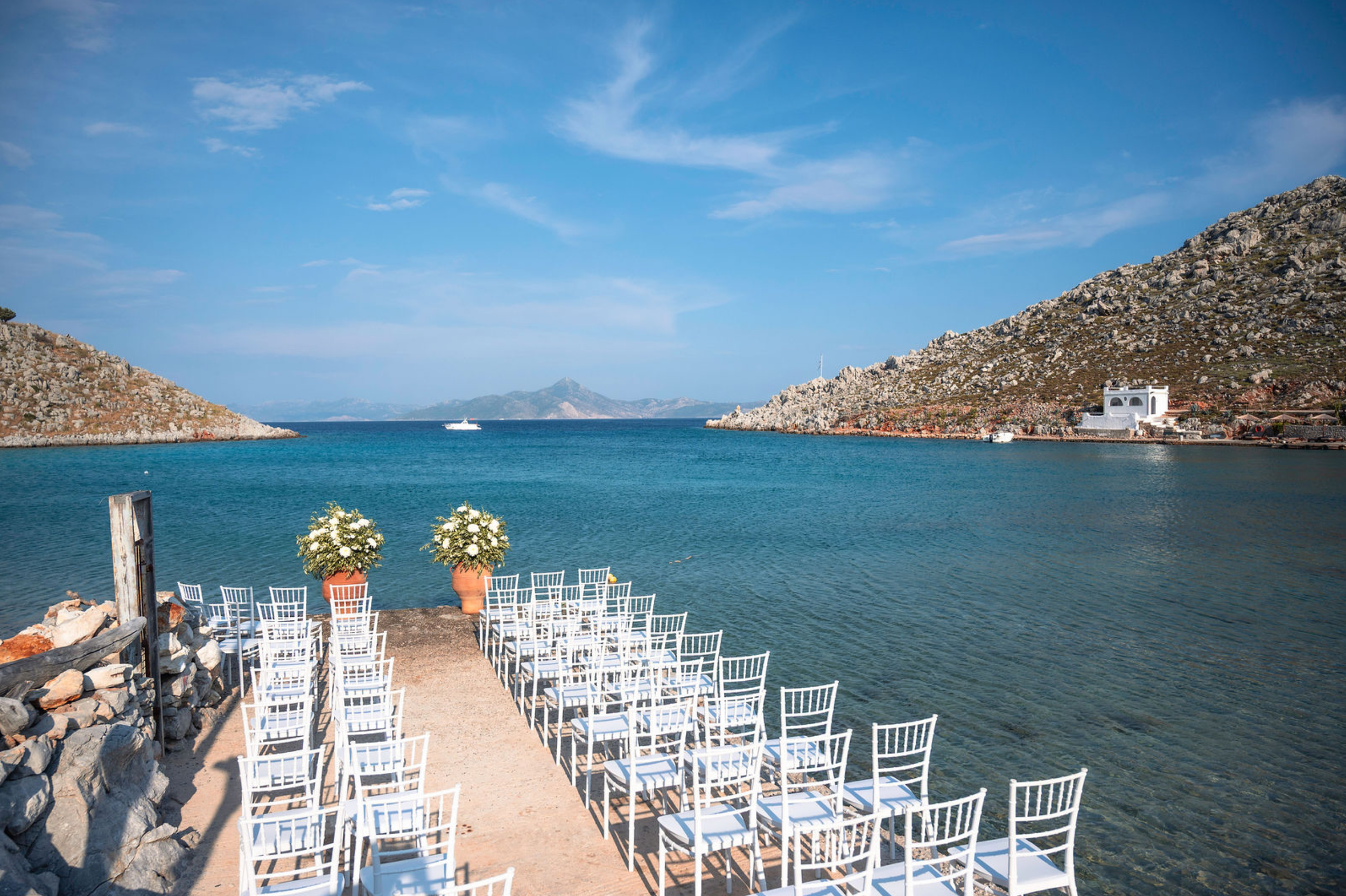 Outdoor wedding setup near a calm blue body of water with rocky hills on both sides and a white building, with chairs arranged in rows and flower pots in the foreground.