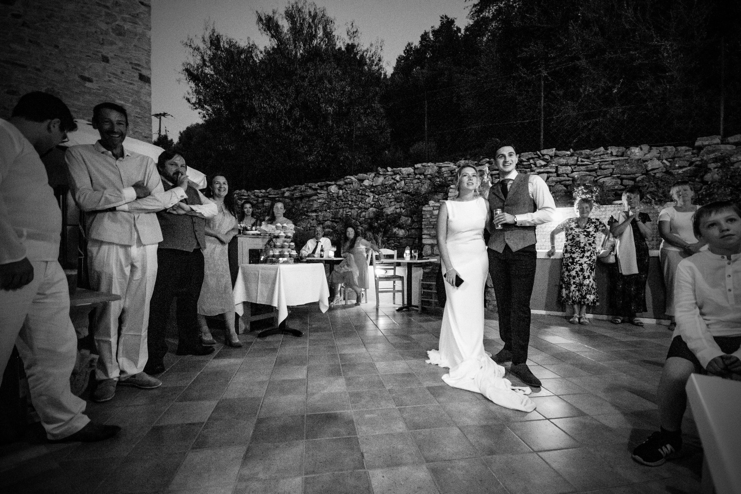 A black and white photo of a wedding reception showing a bride and groom standing together, surrounded by guests, with some smiling, in an outdoor setting at night.