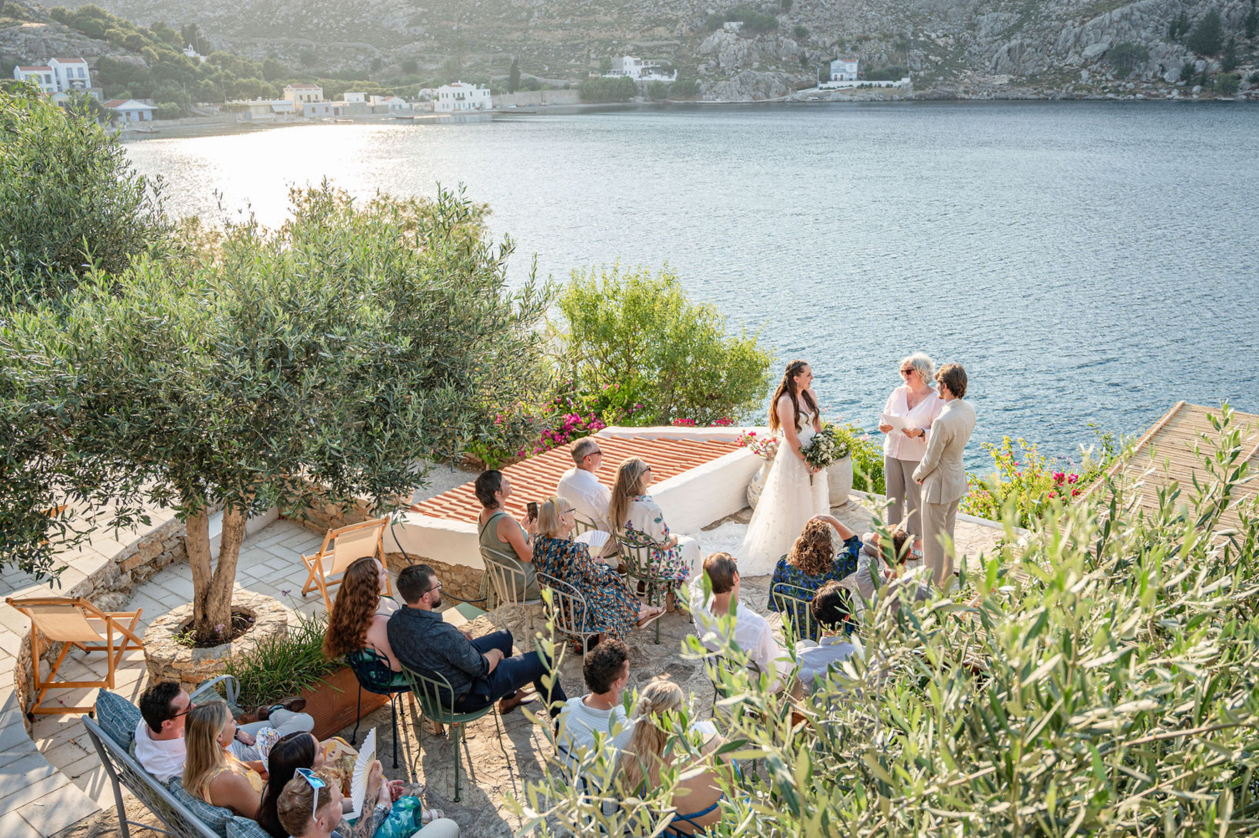 A wedding ceremony taking place outdoors near a body of water with white buildings on a hillside in the background. A bride and groom stand together in front of seated guests, with a woman officiating.