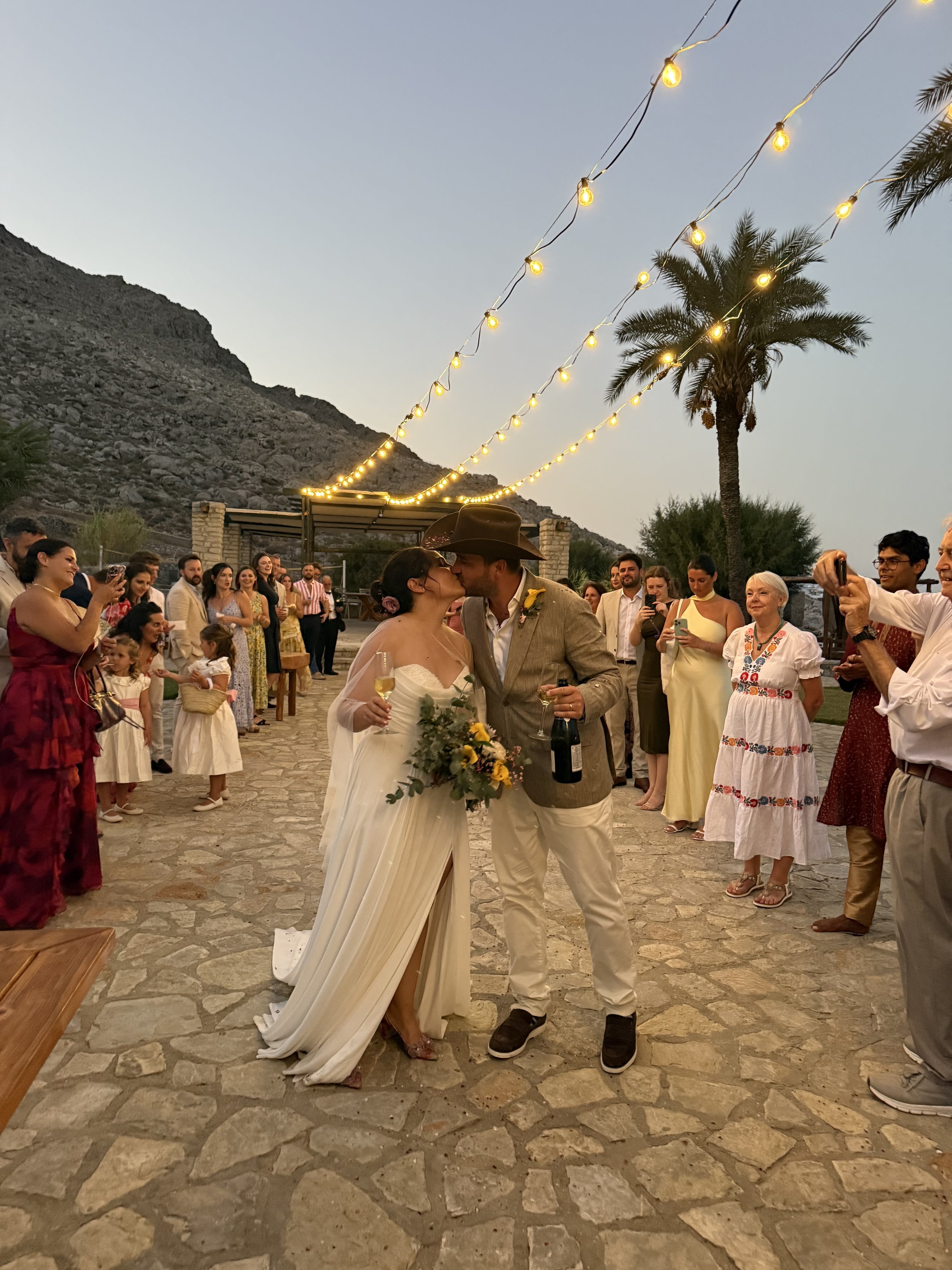 A wedding couple sharing a kiss with guests surrounding them during an outdoor celebration at dusk, decorated with string lights and a palm tree in the background.