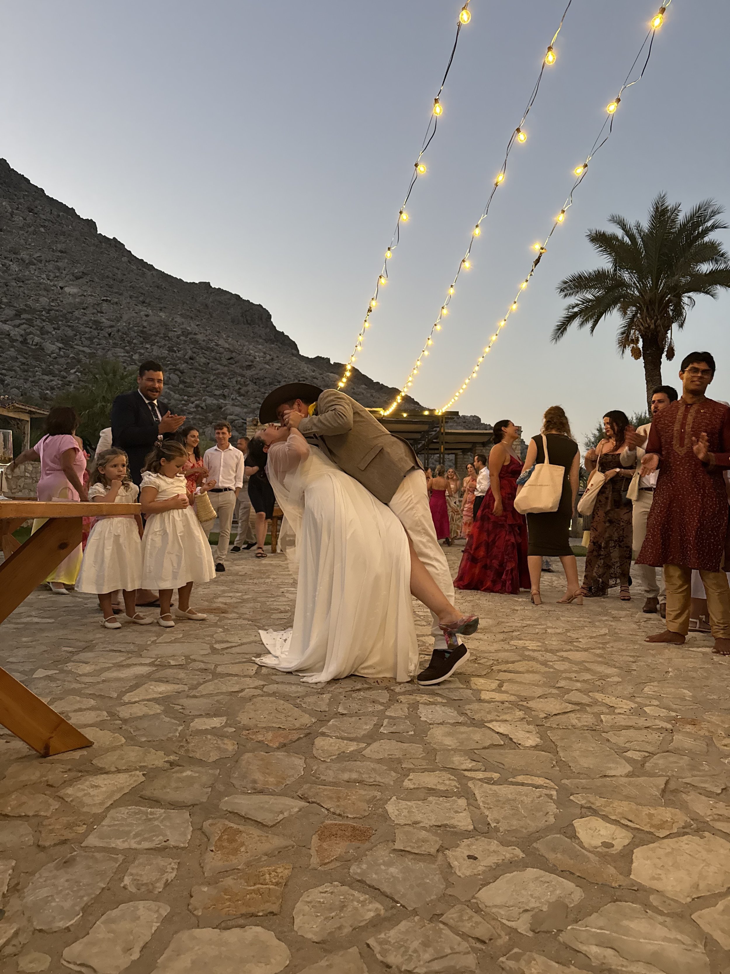 A bride and groom kissing at their wedding reception outdoors during dusk, surrounded by guests, with string lights and a mountain in the background.