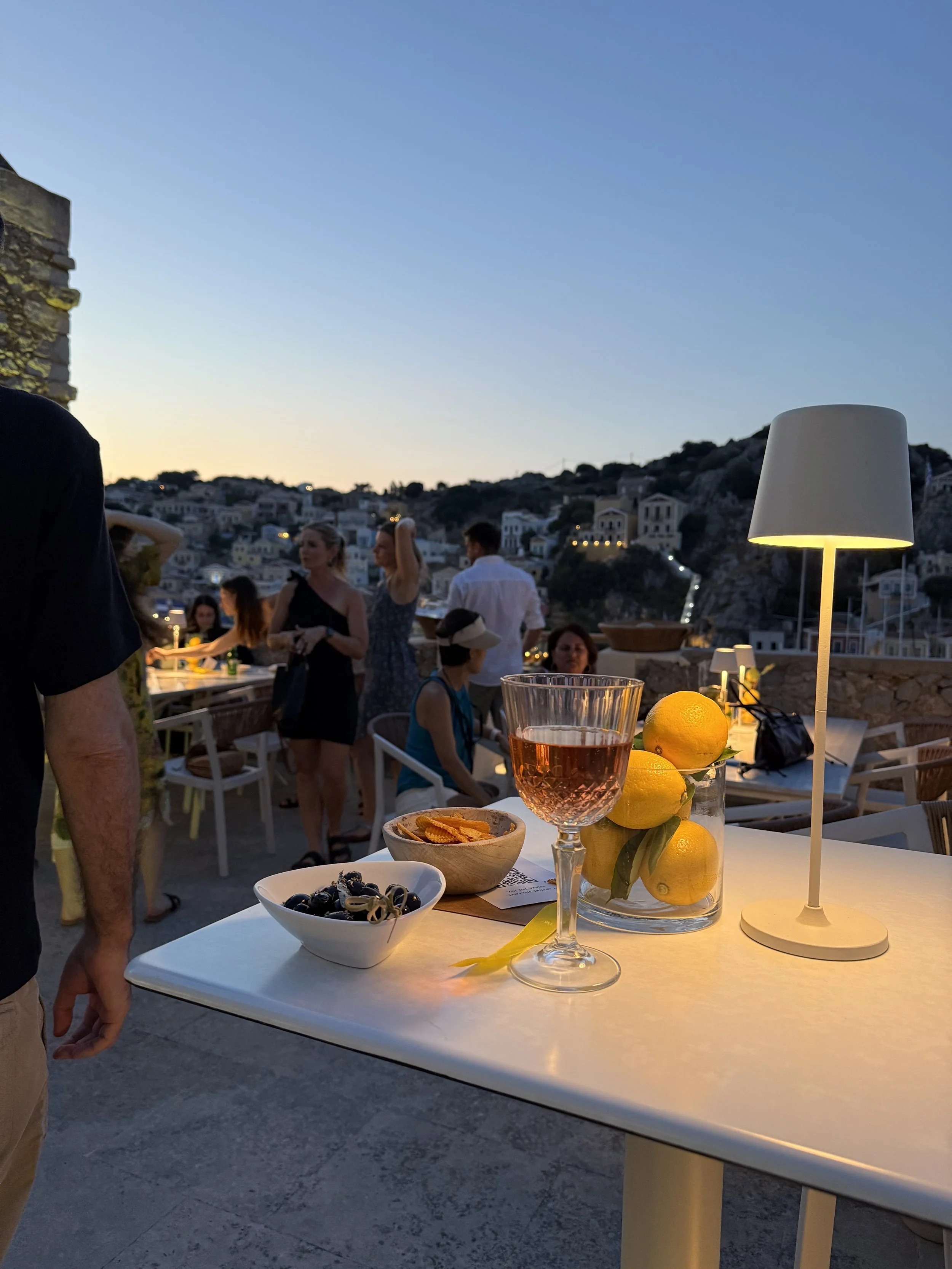 An outdoor gathering at sunset showing a white table with a drink, lemons, and snacks, surrounded by people socializing with a hillside town in the background.