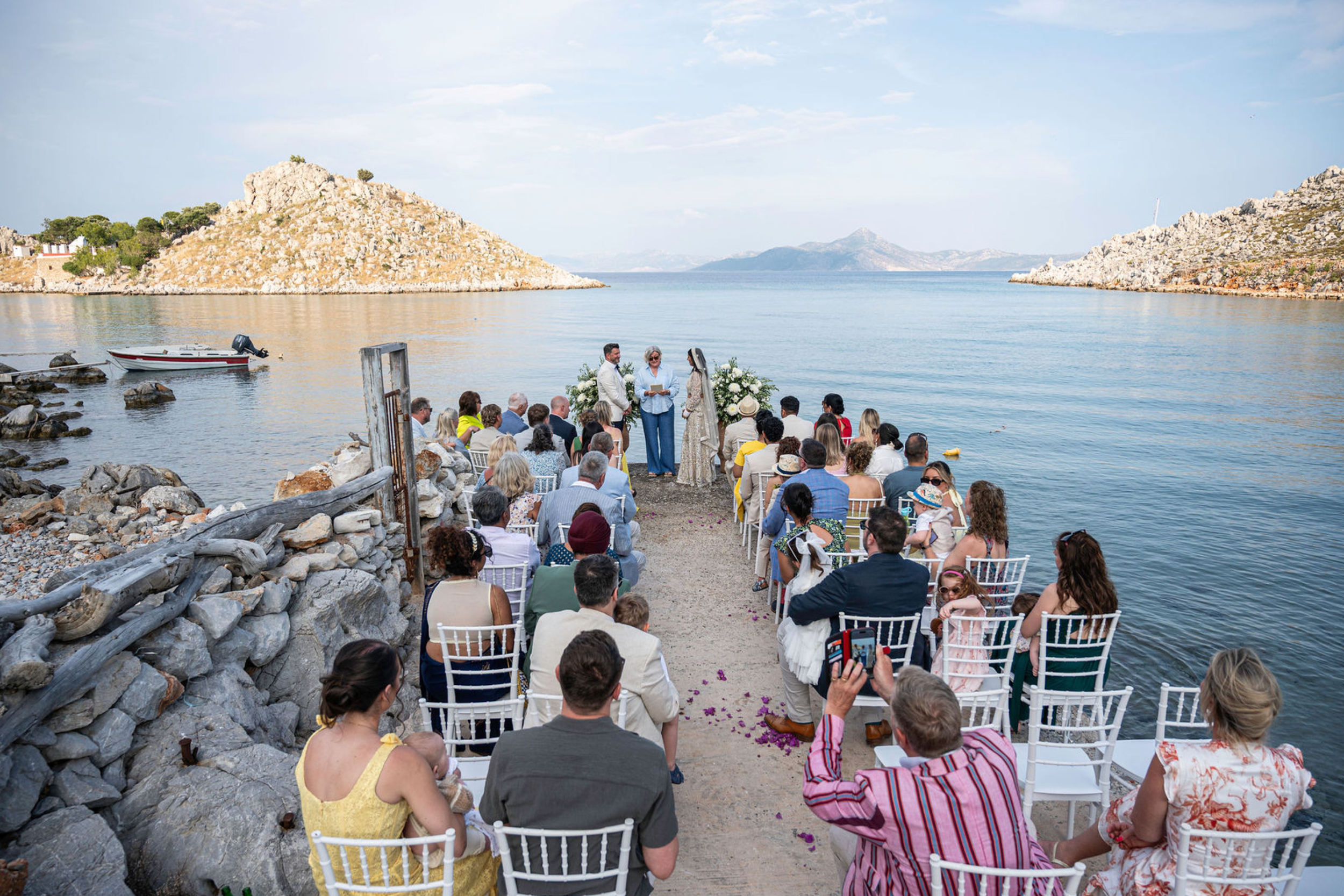 A wedding ceremony held outdoors by a body of water, with guests seated on white chairs and a couple standing with an officiant, surrounded by flowers and scenic rocky hills in the background.