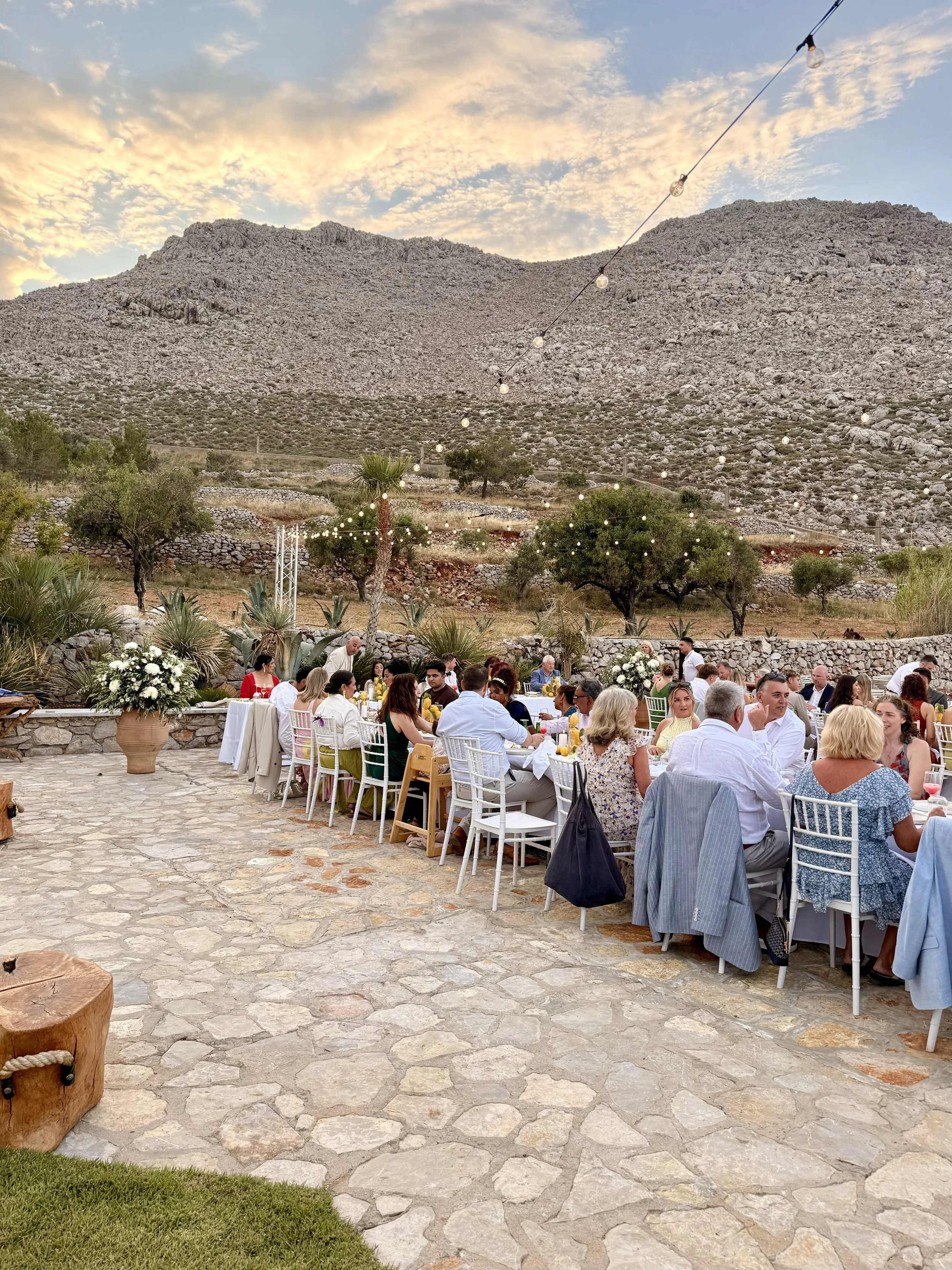An outdoor gathering with a large group of people seated at tables decorated with white tablecloths and floral centerpieces, set against a backdrop of rocky mountains and a colorful sky.