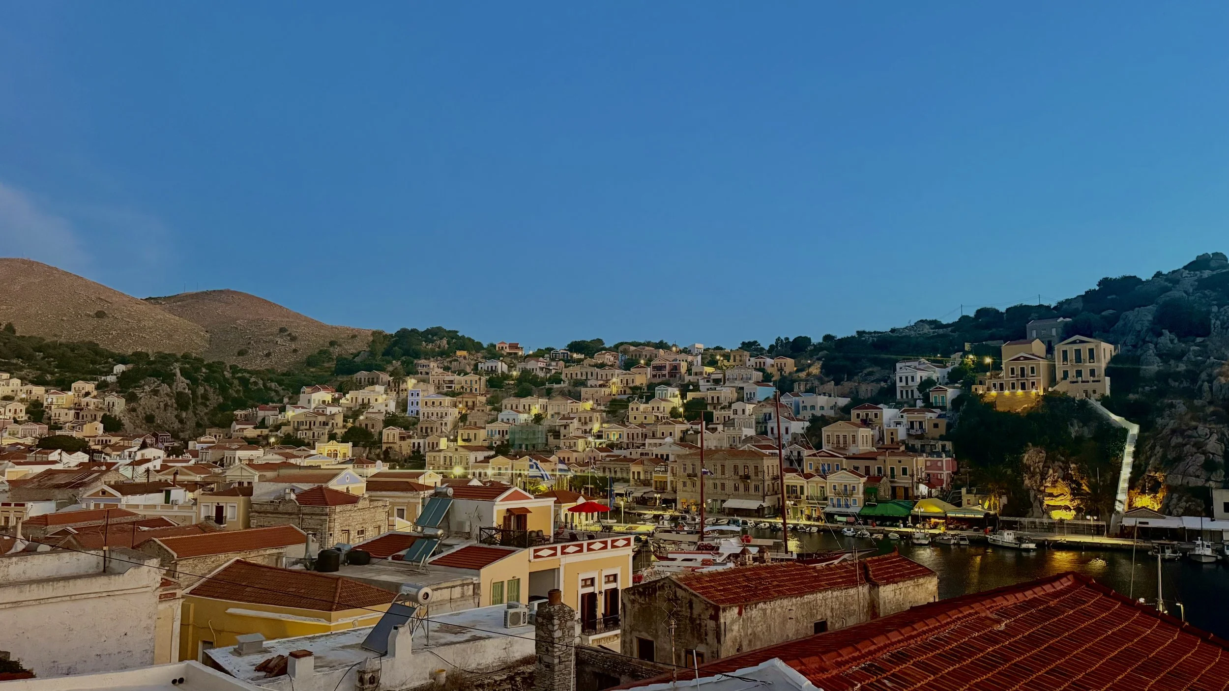 Coastal town with colorful houses on hillside, boats in marina, and hills in background during dusk with clear sky.
