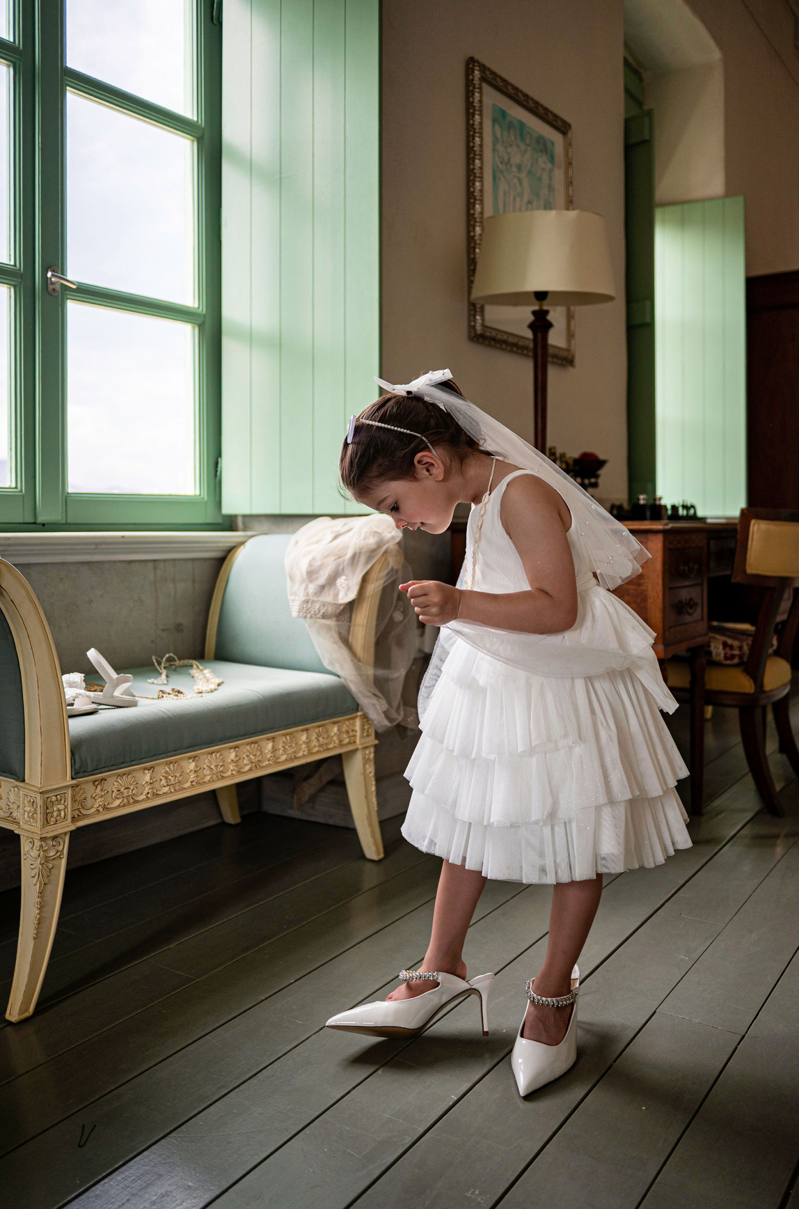 A young girl in a white dress and veil, likely a flower girl, putting on white high heel shoes in a room with green shutters and vintage furniture.
