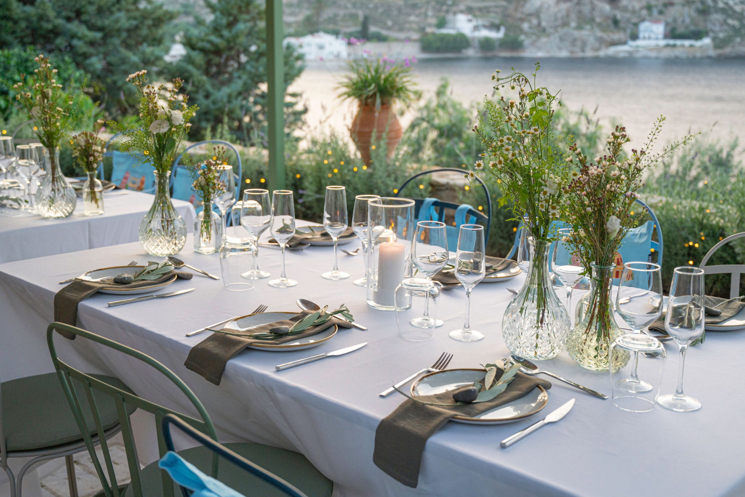 A table set for a meal outdoors with white tablecloth, glassware, silverware, small flower arrangements in glass vases, and a lit candle. In the background, there is a river, greenery, and houses on a hillside.