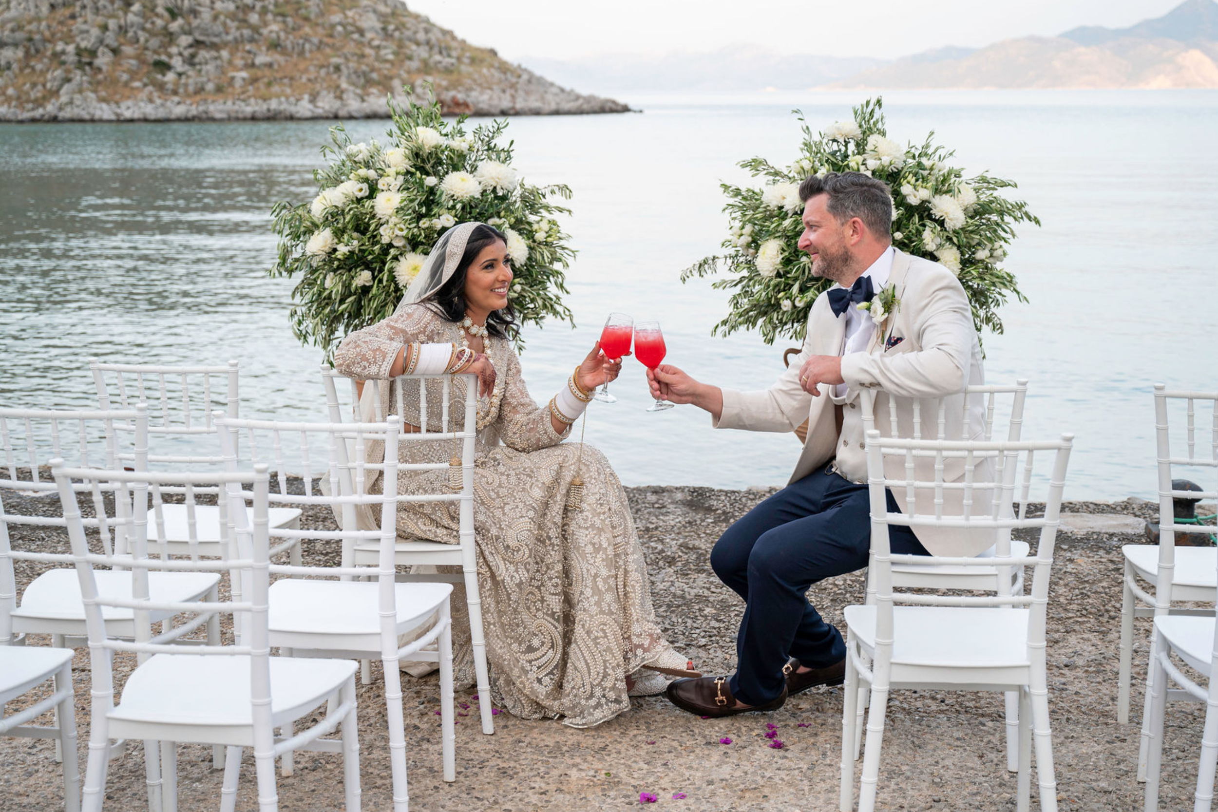 A bride and groom cheer with red beverages at their outdoor wedding ceremony near a lake, with white chairs and floral arrangements behind them.
