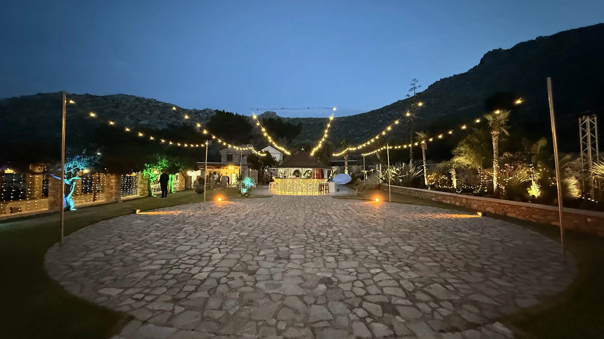Outdoor event space at dusk with string lights, decorative trees, and a stone paved walkway leading to a pavilion in a mountainous area.