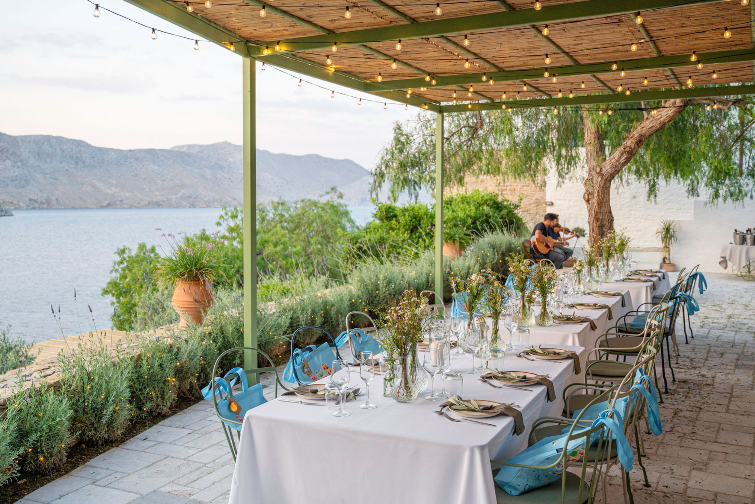 An outdoor dining setup on a patio overlooking a lake with mountains in the background. The patio is decorated with string lights, potted plants, and has a long table with white tablecloths, glassware, and floral centerpieces. There are two musicians