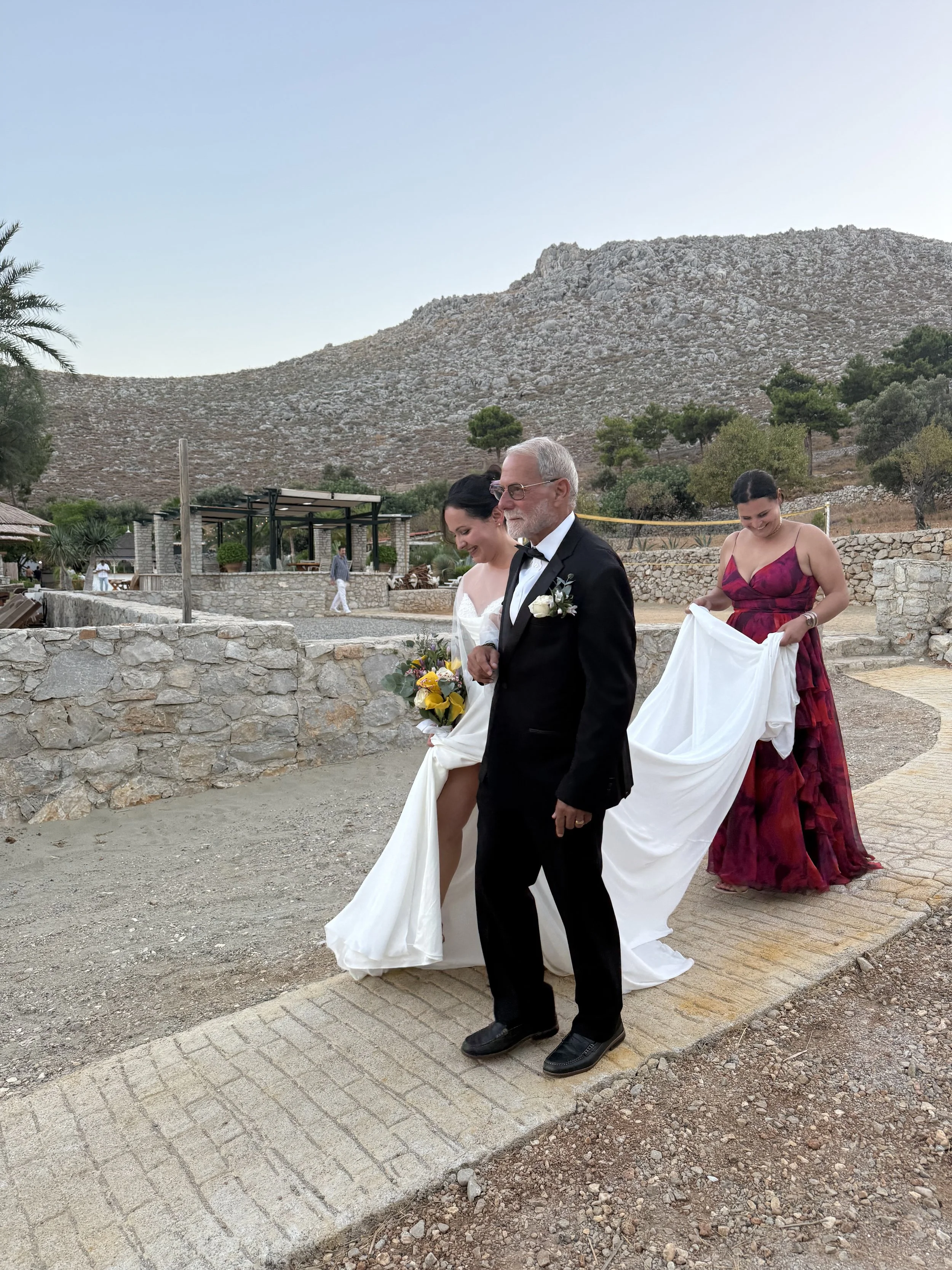 A wedding scene featuring an older man in a tuxedo with a white flower boutonniere walking next to a woman in a white wedding dress holding a bouquet of flowers. Behind them, a woman in a red and purple dress is holding the train of the wedding dress