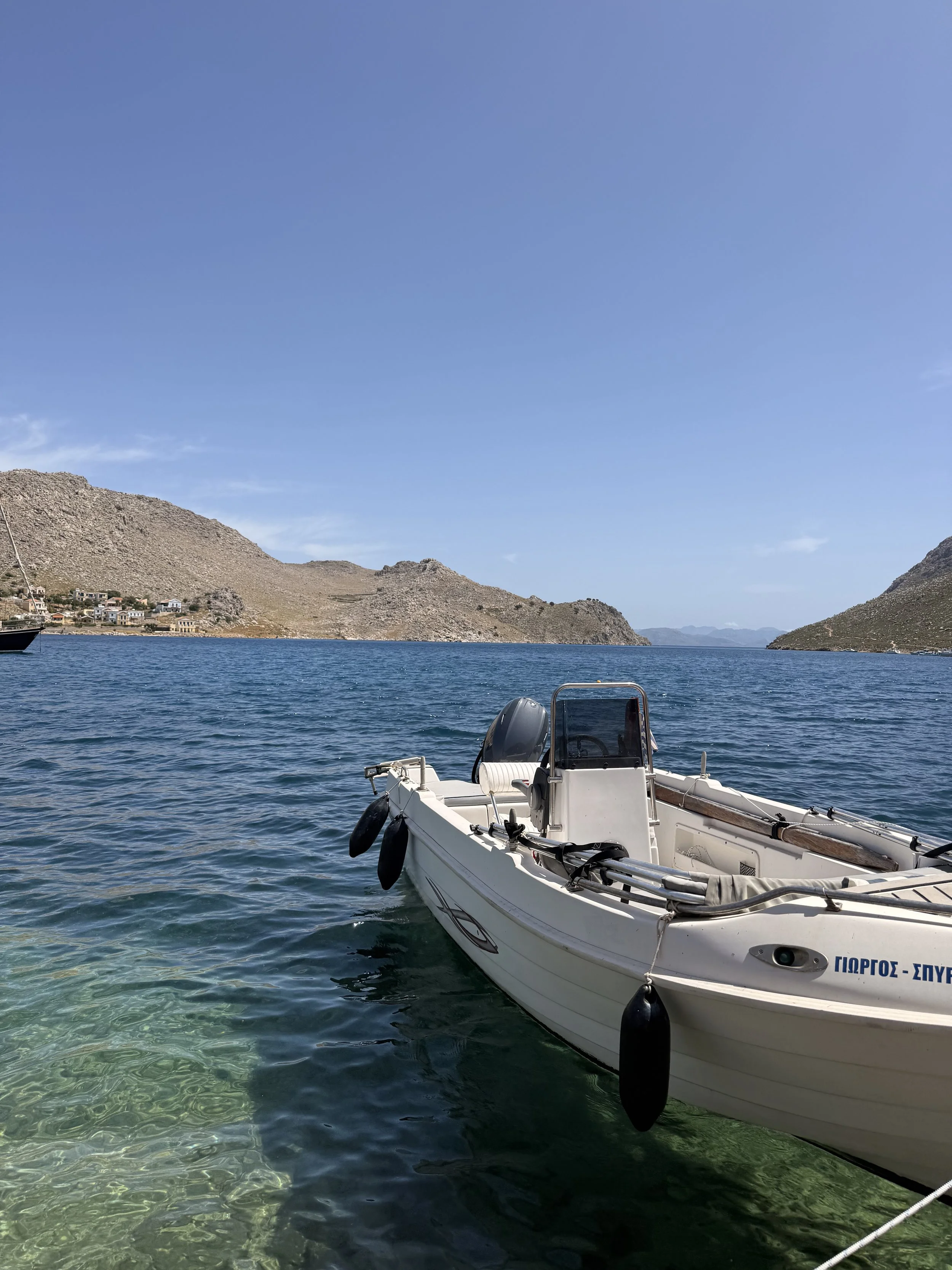 A small white motorboat floating on clear blue water near a rocky shoreline with a small village and hills in the background under a blue sky.