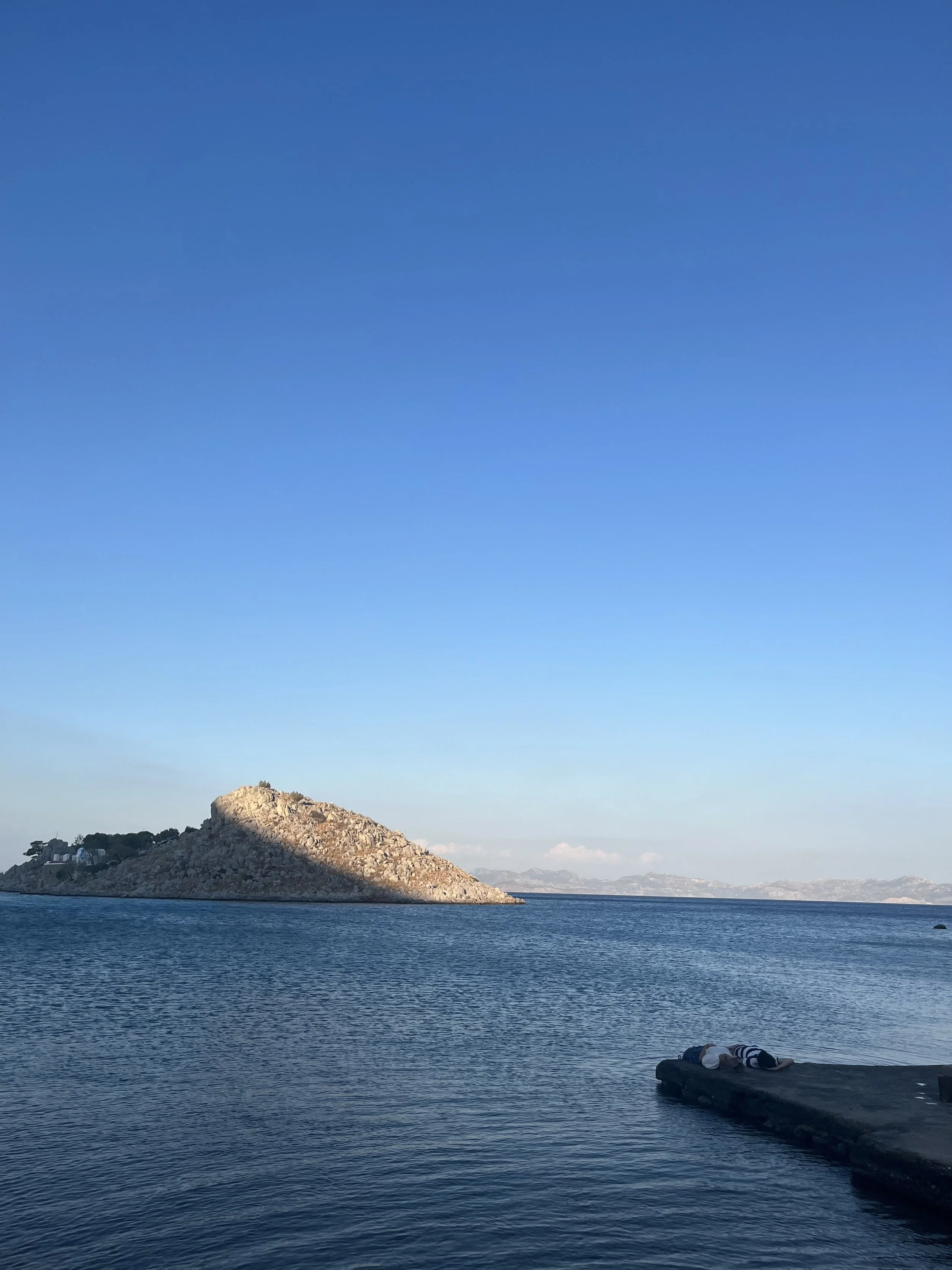A coastal scene with a body of water in the foreground, a rocky island or hill on the left, and a clear blue sky overhead.