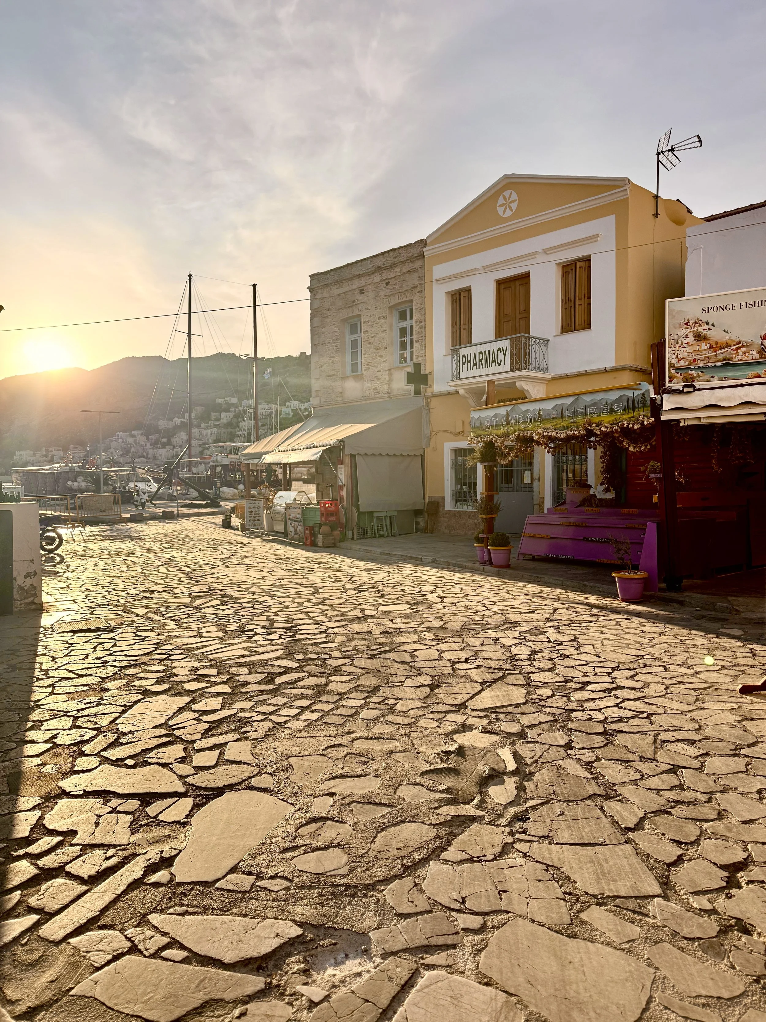 A seaside street at sunset with a cobblestone path, yellow and white buildings, a marina with sailboats, and mountains in the background.