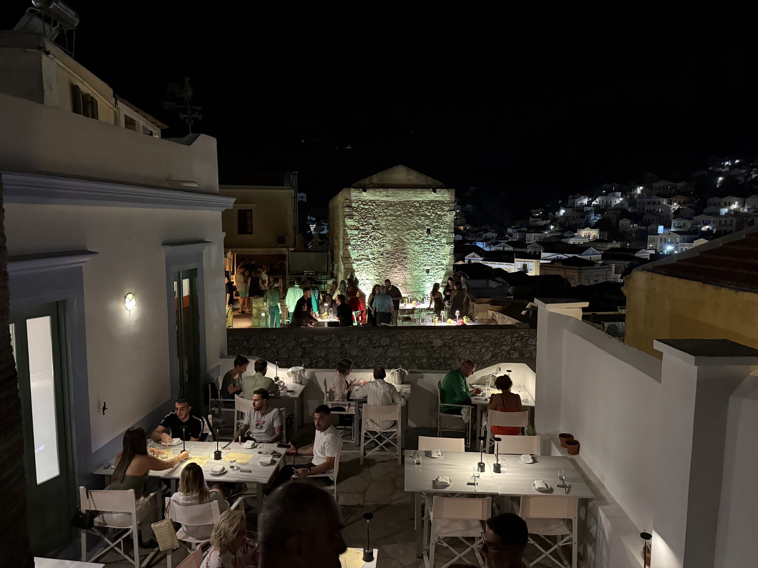 Nighttime scene of a restaurant patio with people dining at tables, illuminated by wall-mounted lights, overlooking a hillside with houses and a historic stone tower.