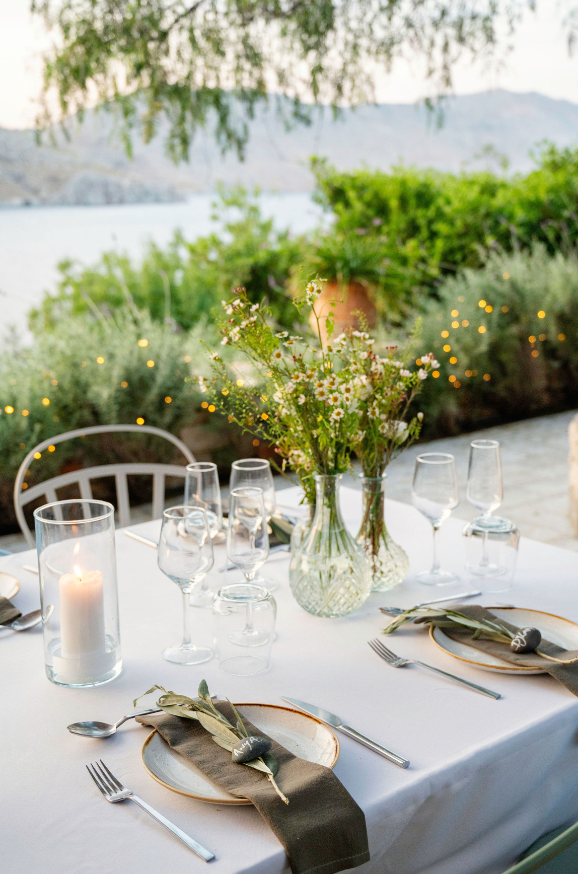 Elegant outdoor dining table set with glasses, plates, silverware, a candle, and a floral centerpiece near a body of water with mountains in the background.