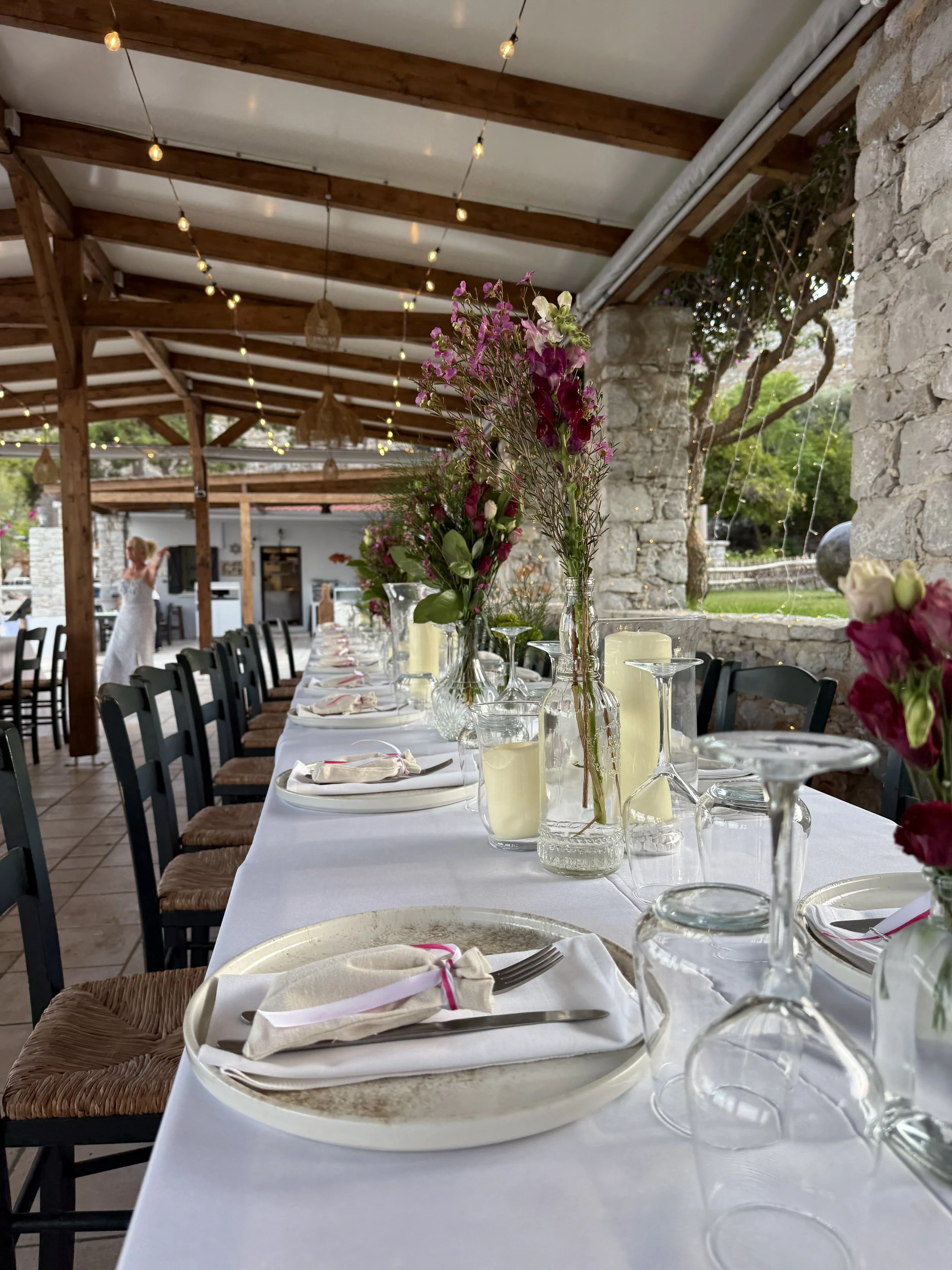 A long dining table set with white tablecloths, pink and purple flower centerpieces in vases, candles, and place settings with napkins and silverware in a rustic outdoor space with wooden beams, string lights, and stone walls.