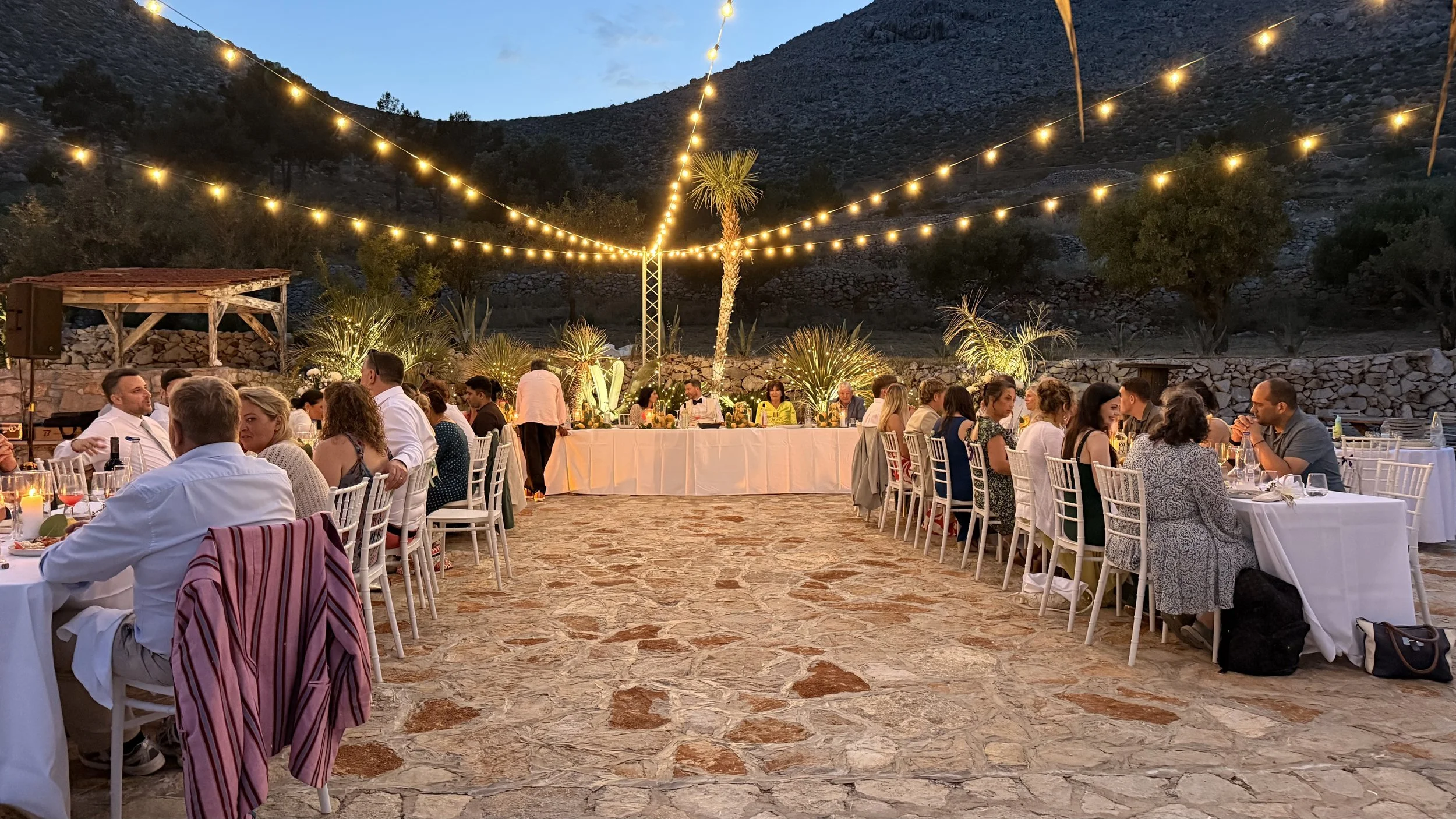 Outdoor evening party with guests seated at round tables under string lights, mountainous backdrop, and a head table with floral arrangements.