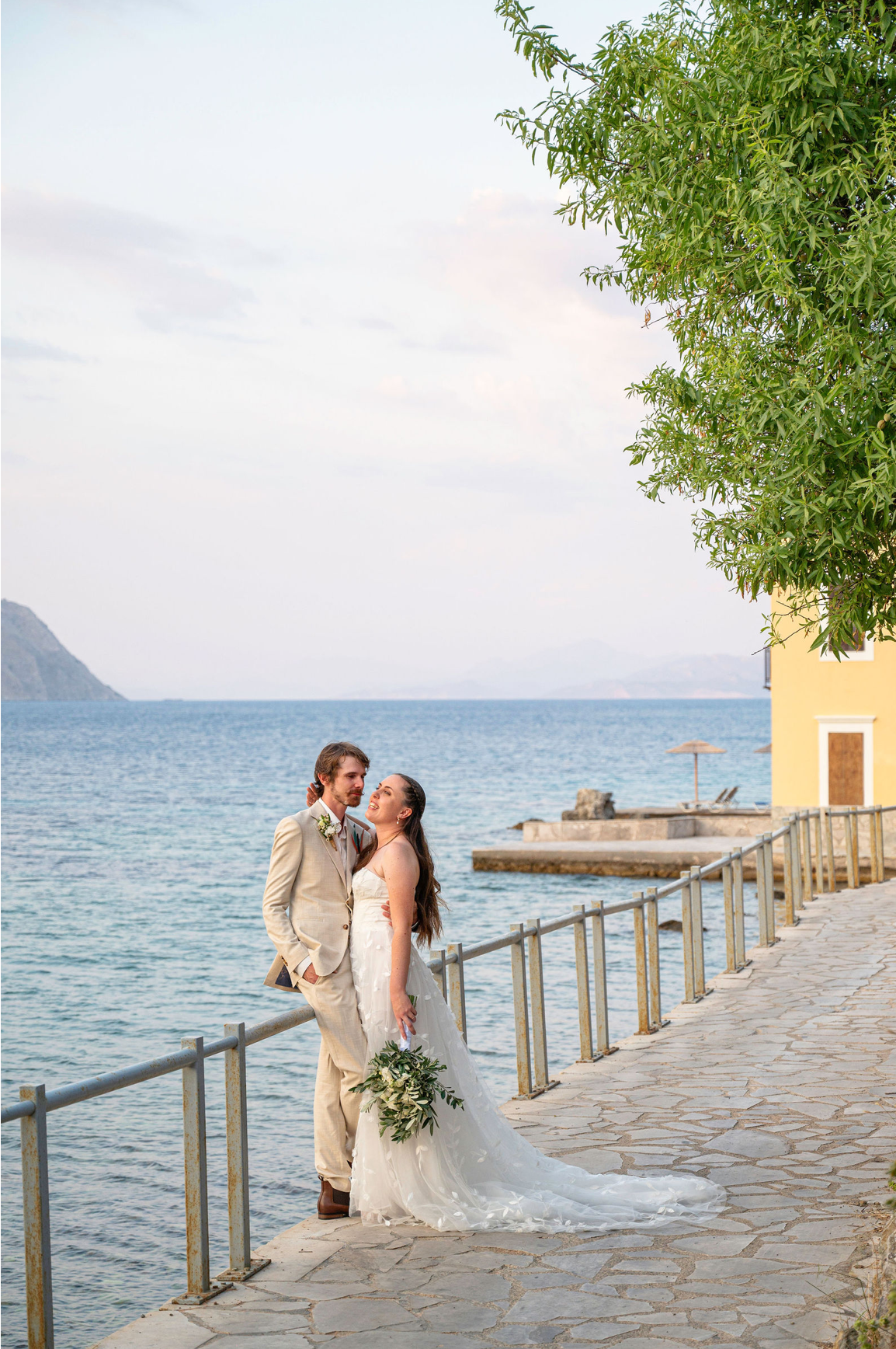 Bride and groom standing by the water on a stone pathway, with a tree and a yellow building in the background.