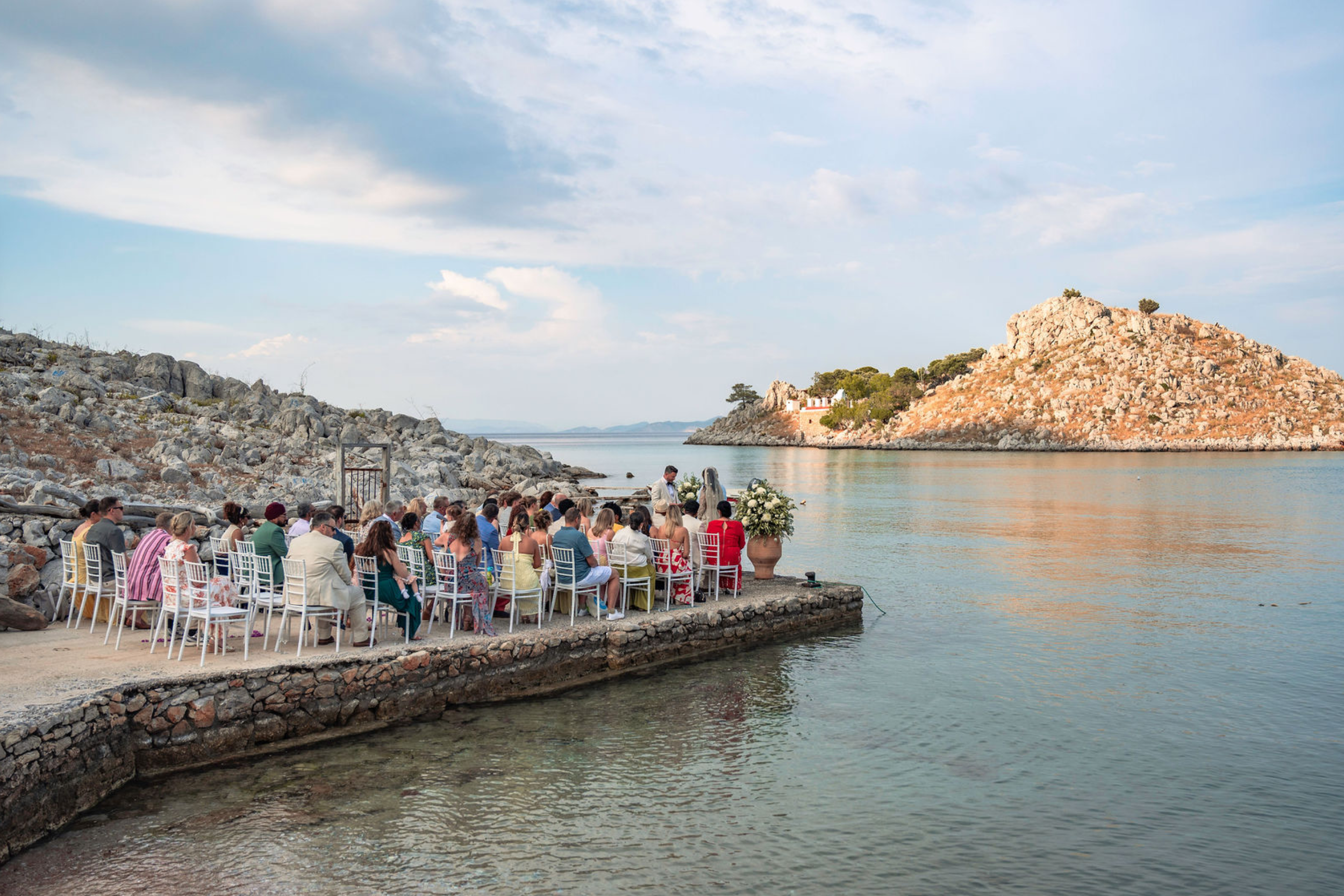 People seated in chairs near water, with a rocky hillside and trees in the background, some clouds in the sky.