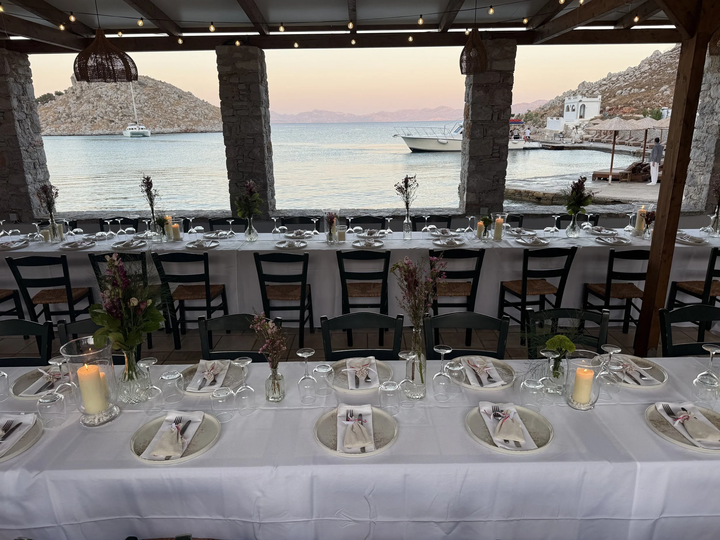 A seaside dining area with a long table set with white tablecloth, candles, flower arrangements, and multiple place settings, overlooking a calm body of water with boats and rocky hills in the background.