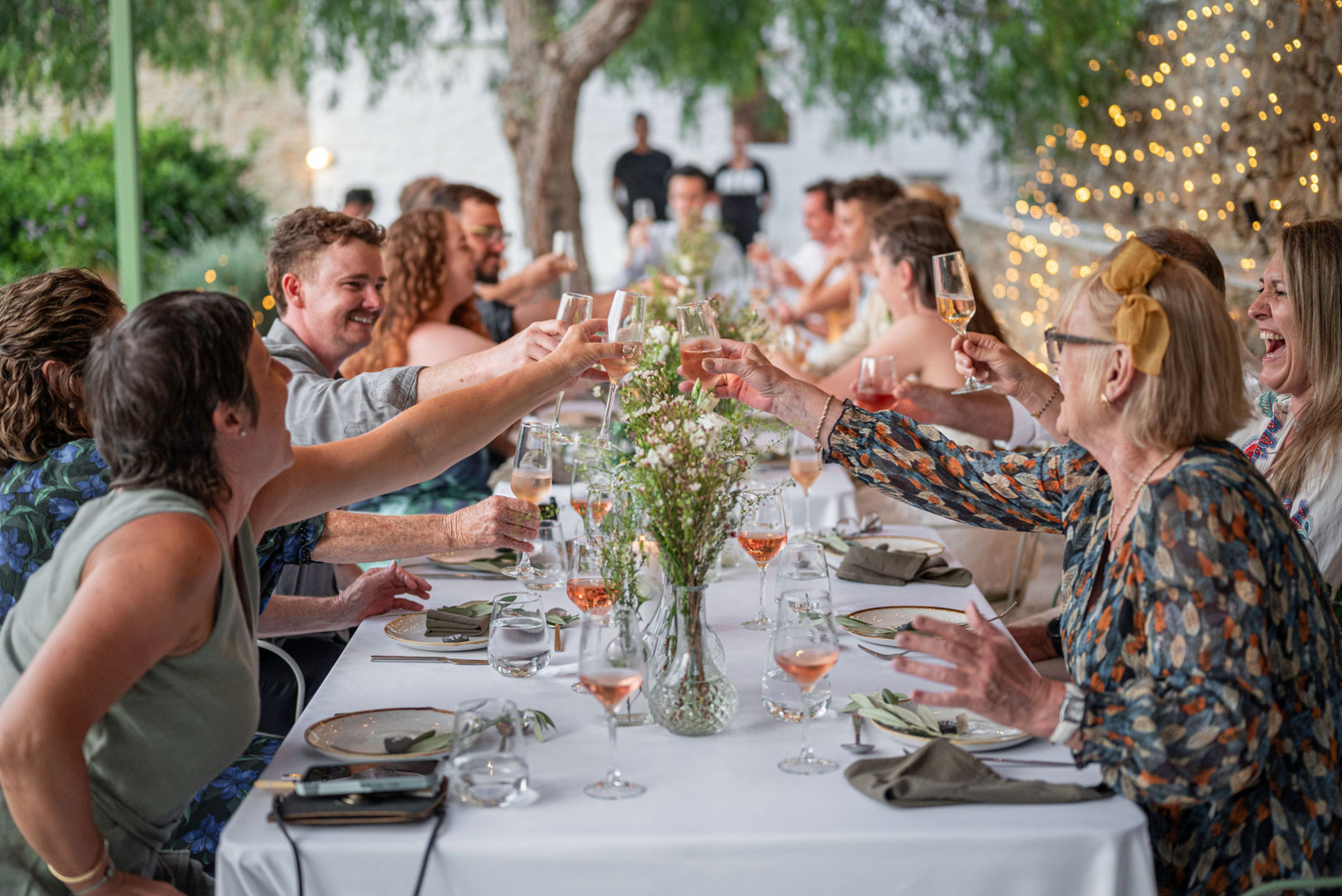 People sitting at a long decorated table outdoors, raising glasses in a toast, with string lights on a tree in the background for a celebration or gathering.