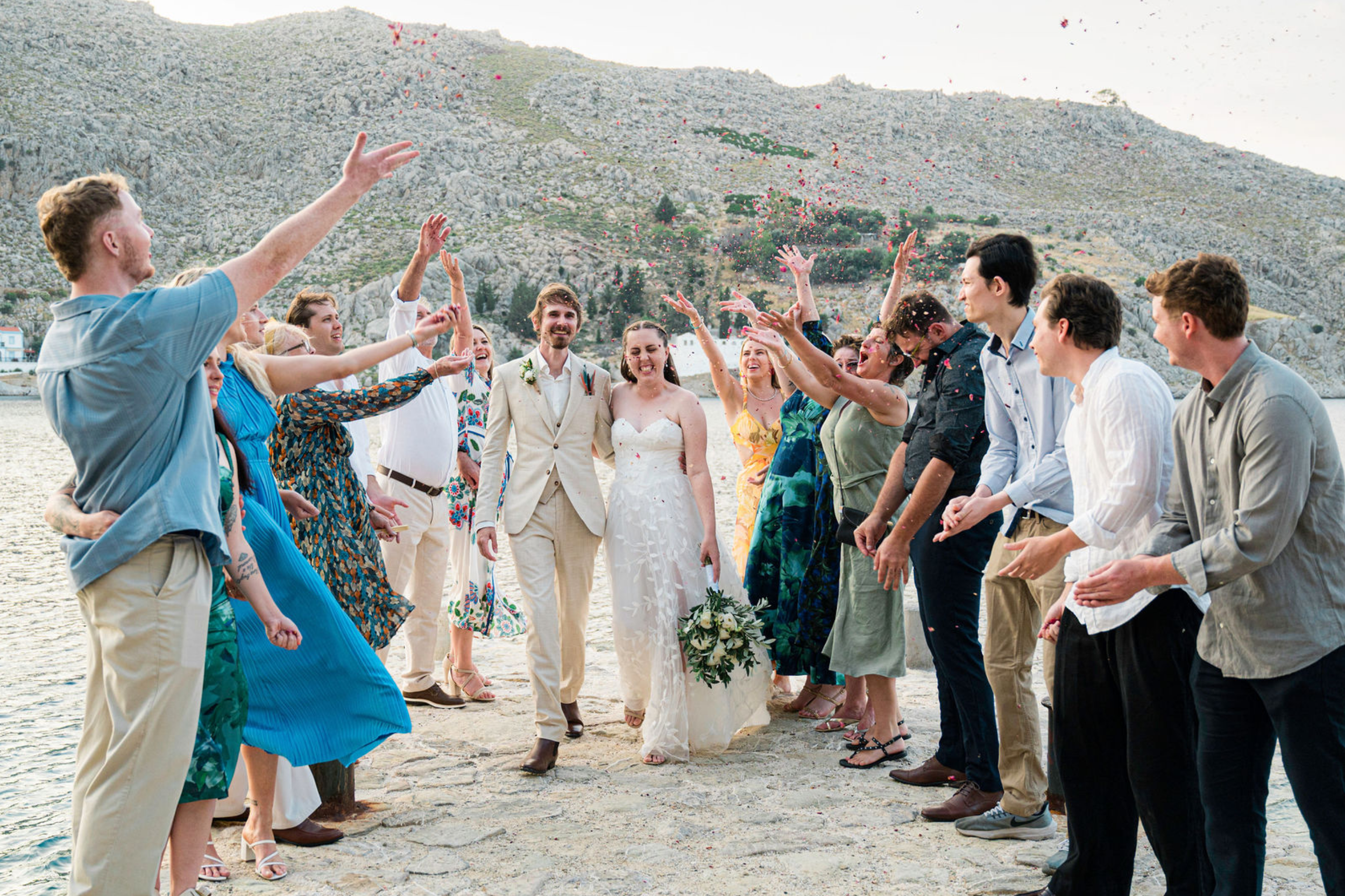 Group of people celebrating a wedding on the beach, with a bride and groom at the center and guests throwing flower petals.