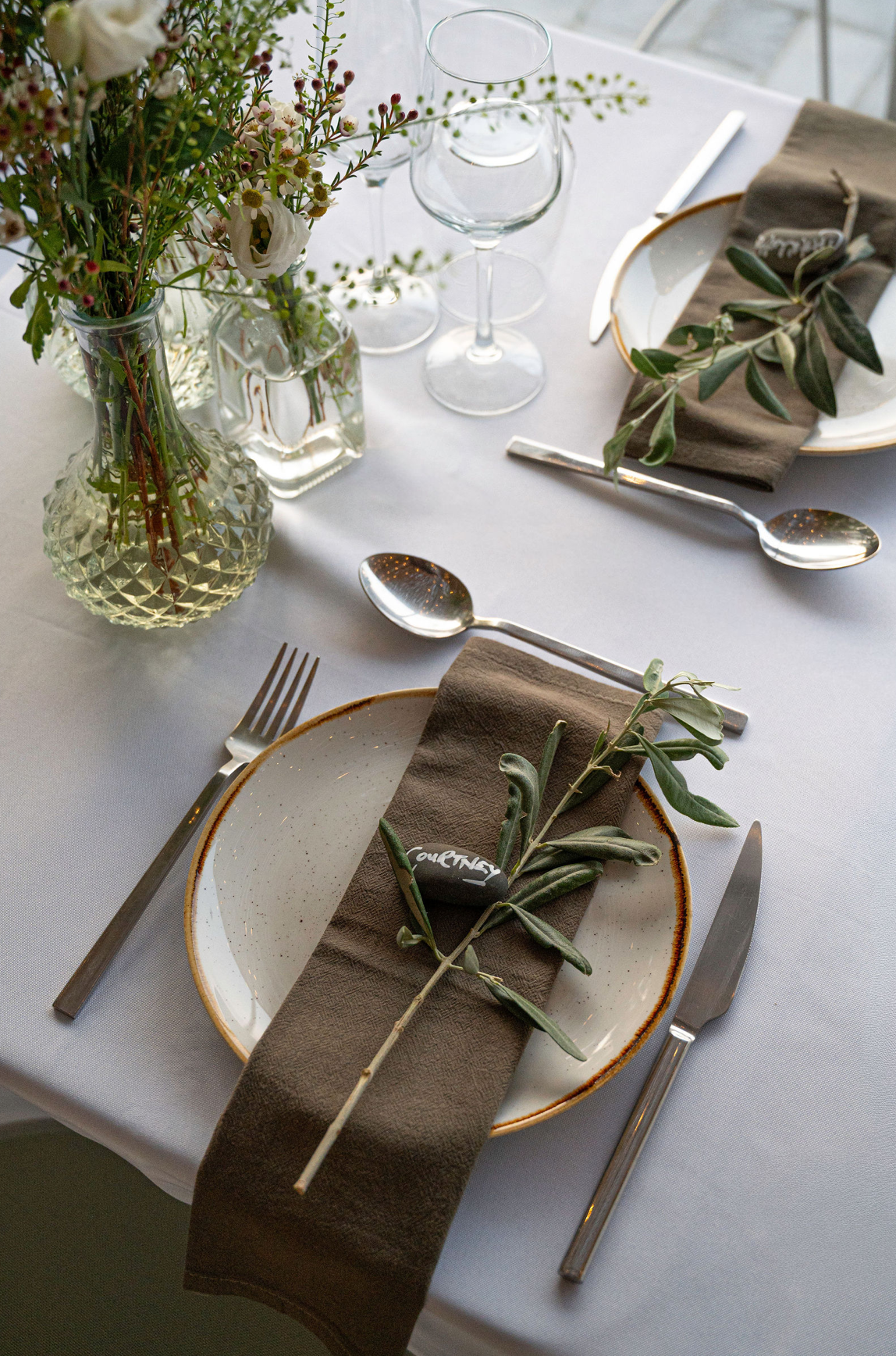 A table setting with a white tablecloth, featuring a white plate with a brown rim, a brown cloth napkin, a sprig of greenery, and a small blackboard with white writing. Silverware includes a fork on the left and a knife on the right. Two glasses are 