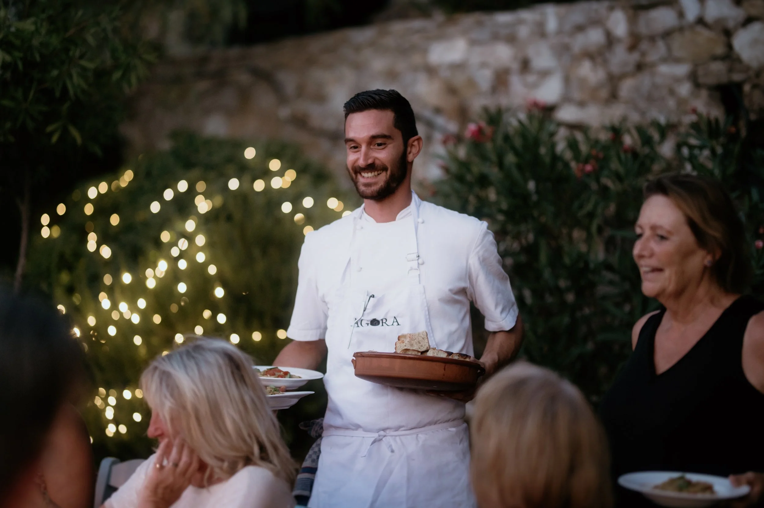 A smiling waiter in a white shirt with an apron serving food to diners at an outdoor restaurant, with fairy lights and greenery in the background.
