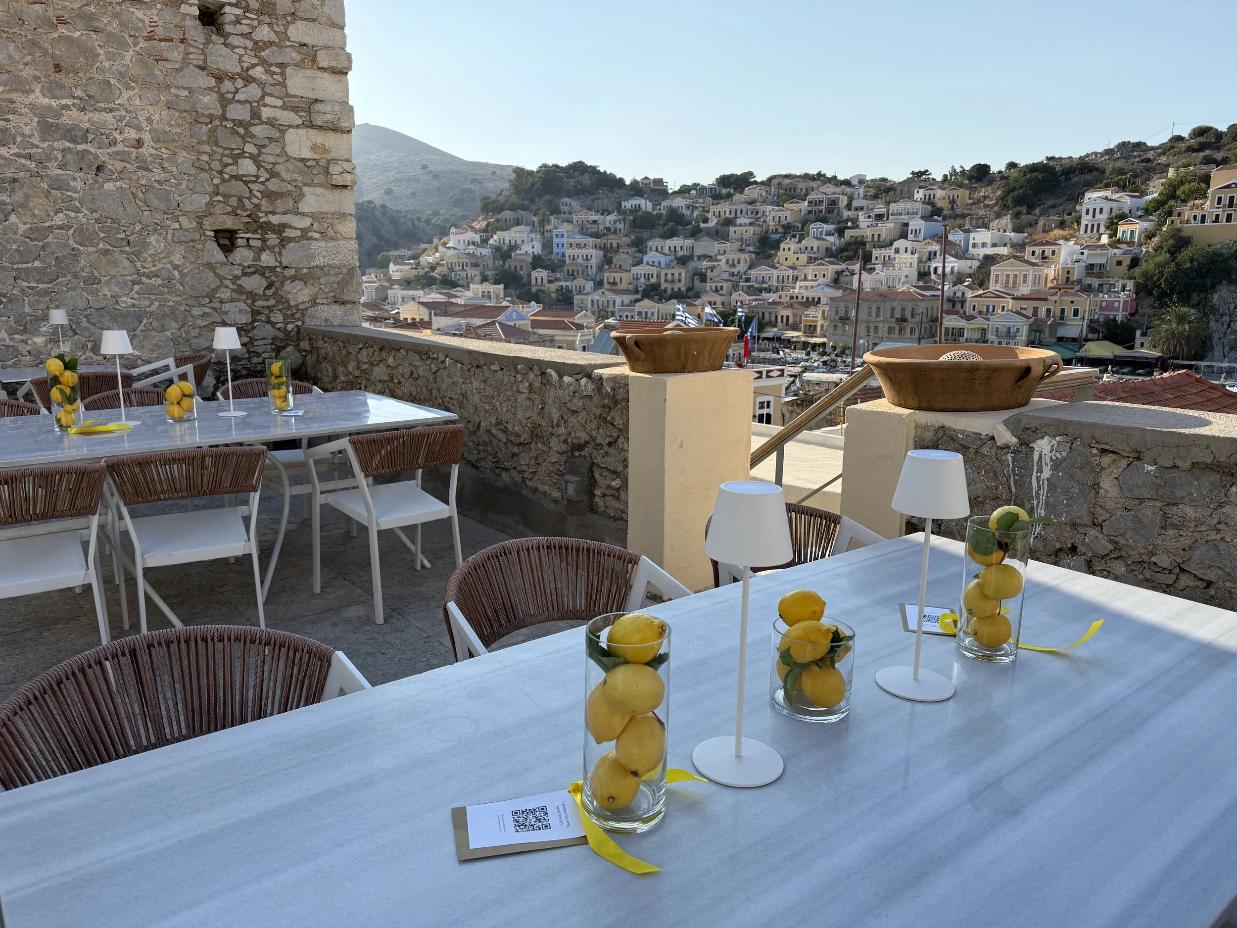 Outdoor dining area with a stone wall, white tables, chairs with woven backs, and decorative glasses filled with lemons and yellow ribbons, overlooking a hillside town with colorful houses and flags.