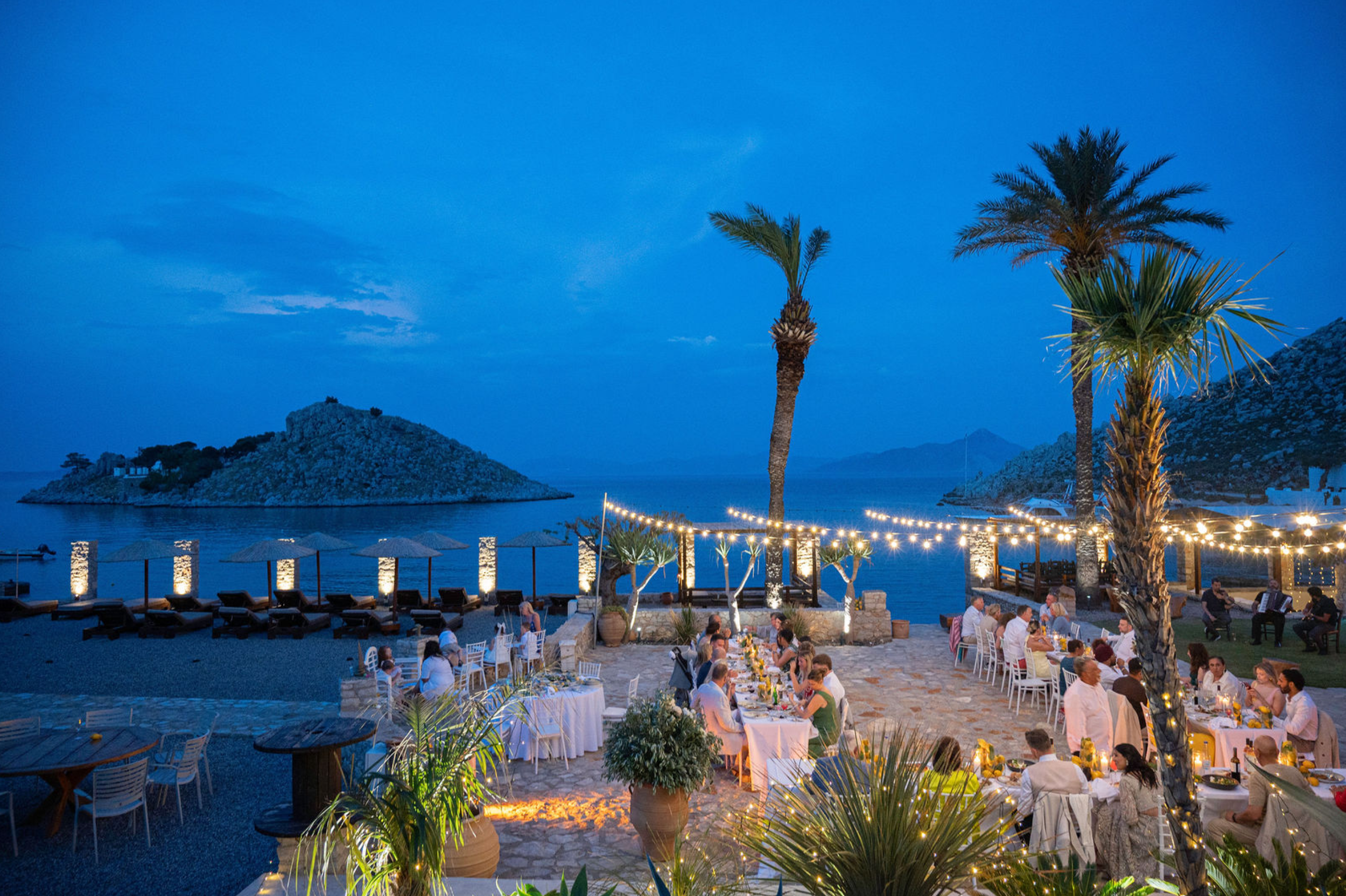 A beachside restaurant during the evening with tables filled with people, palm trees, string lights, and a view of the water and distant islands.