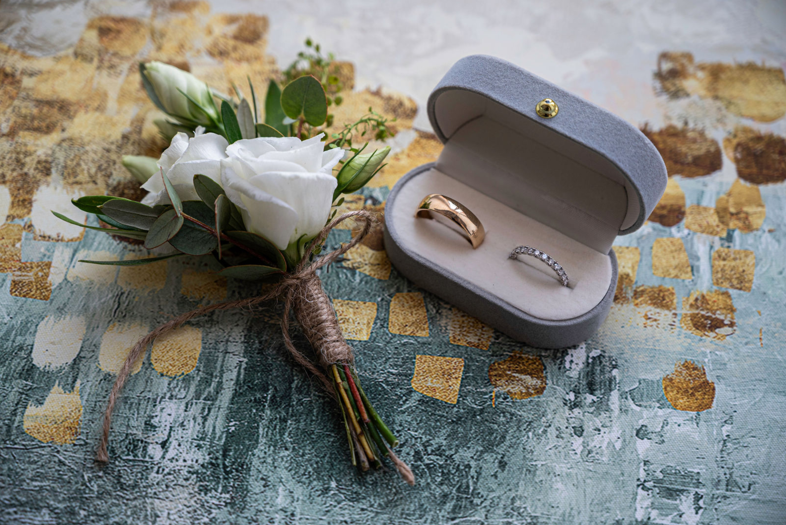 A bouquet of white flowers with green leaves and a tied jute string next to a gray jewelry box with a gold ring and a diamond ring inside. The scene is set on a colorful textured fabric surface.