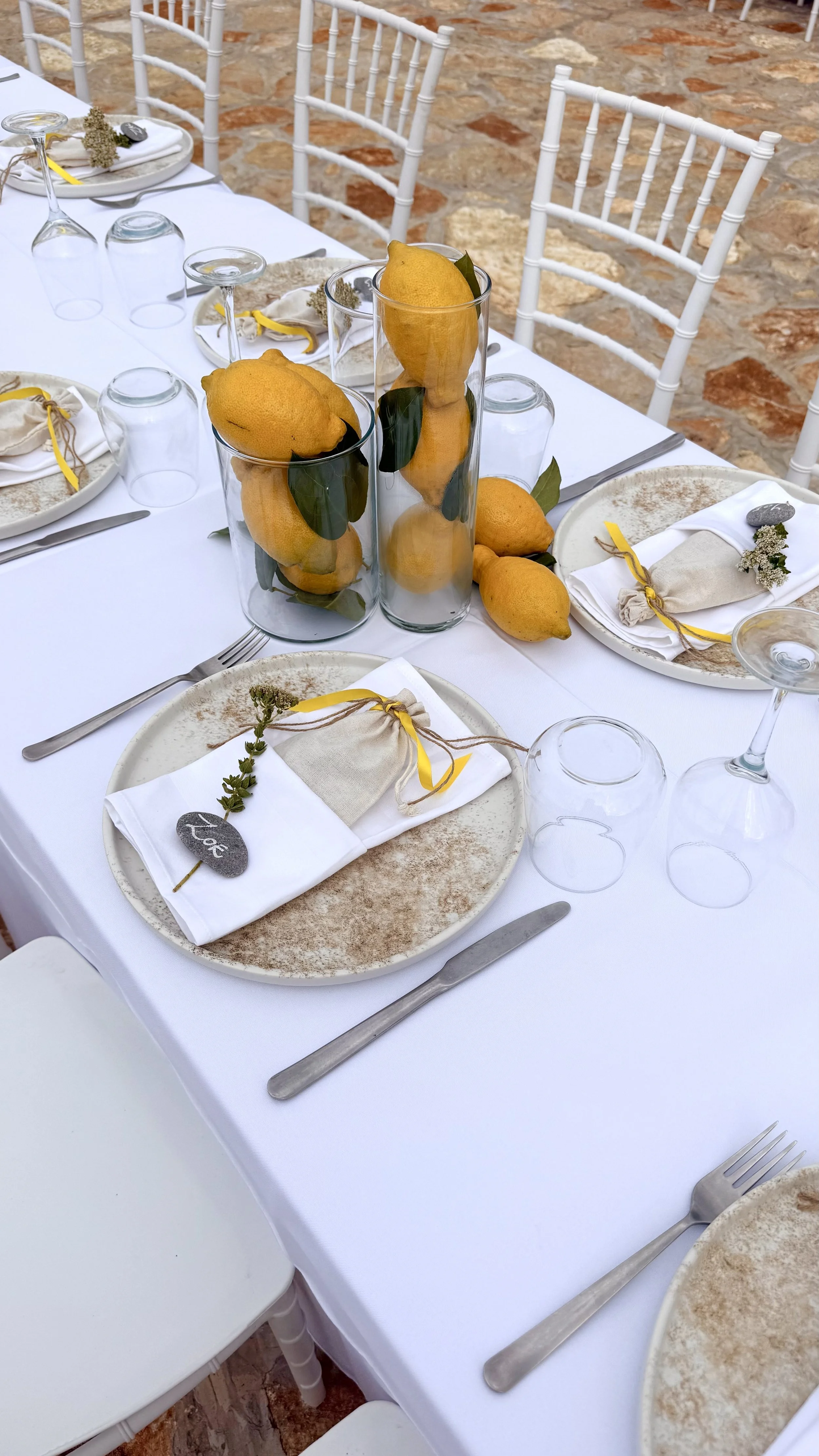 Set dining table with white tablecloth, place settings including plates, napkins with yellow ribbons, silverware, overturned wine glasses, and centerpieces with lemons in tall glass vases, on a rustic brick floor outdoors, surrounded by white chairs.