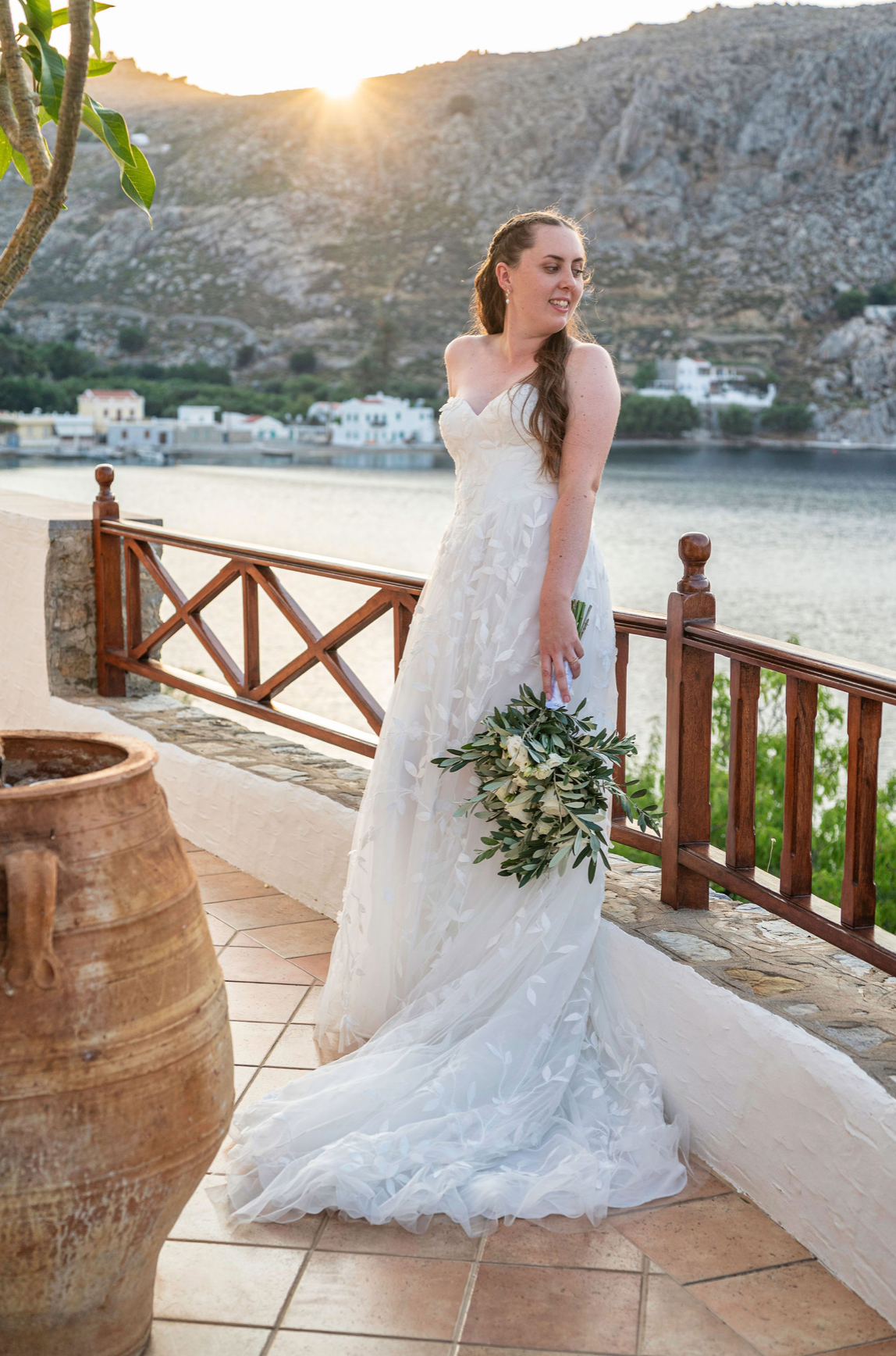 A woman in a white wedding dress holding a bouquet of greenery and white flowers standing on a balcony overlooking water and mountains during sunset.