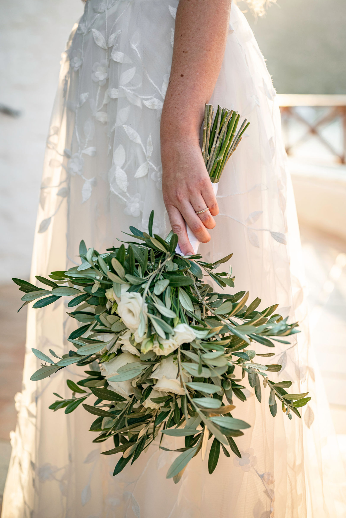 A bride holding a bouquet of white roses and greenery, wearing a white wedding dress with floral embroidery, outdoors during sunset.