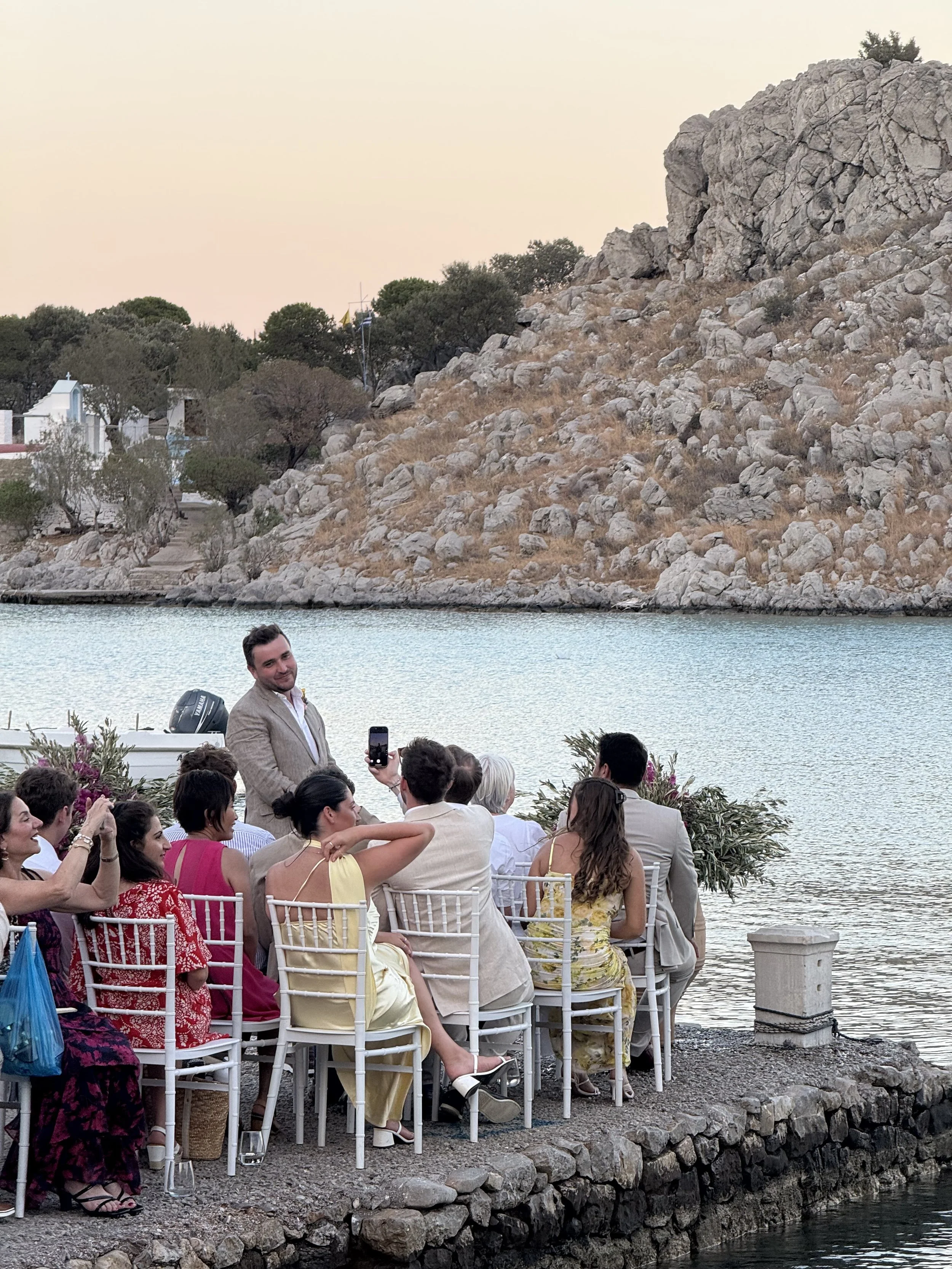 People at an outdoor wedding or event near a water body with a rocky hillside and trees in the background.