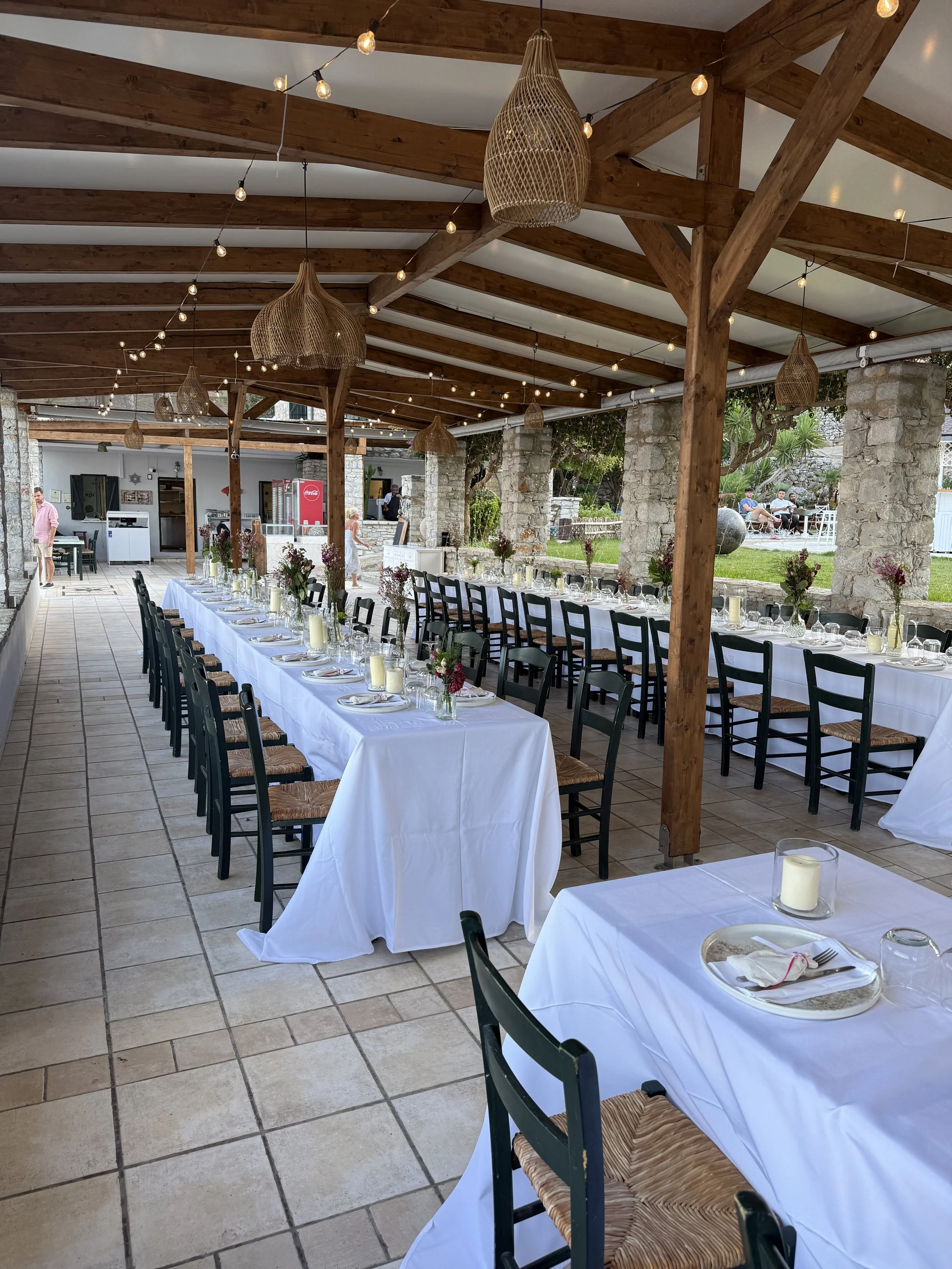 Long rectangular dining table with white tablecloth, set with plates, glasses, and floral centerpieces, under a wooden pergola with rustic hanging lights, in an outdoor event space with stone pillars and a lawn in the background.