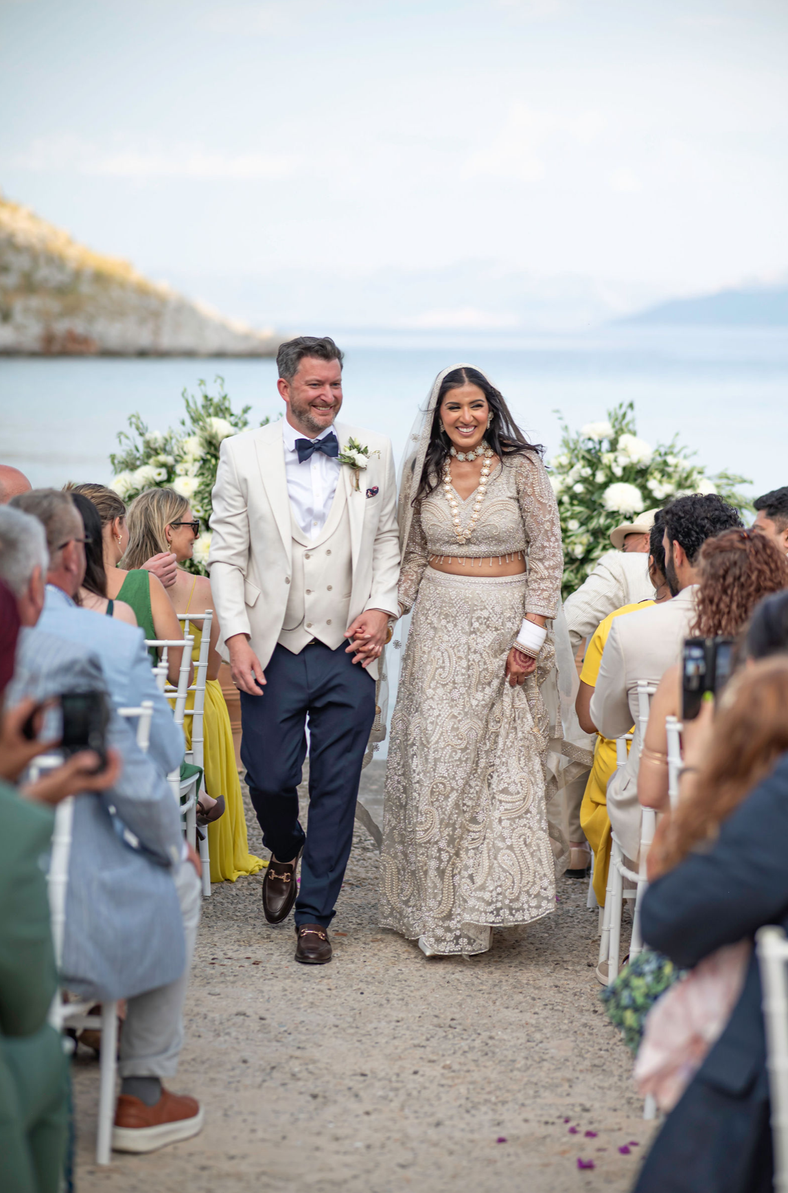 A bride and groom walking hand in hand down the aisle at an outdoor wedding by the water, surrounded by guests seated in white chairs, with a backdrop of rocks and water.