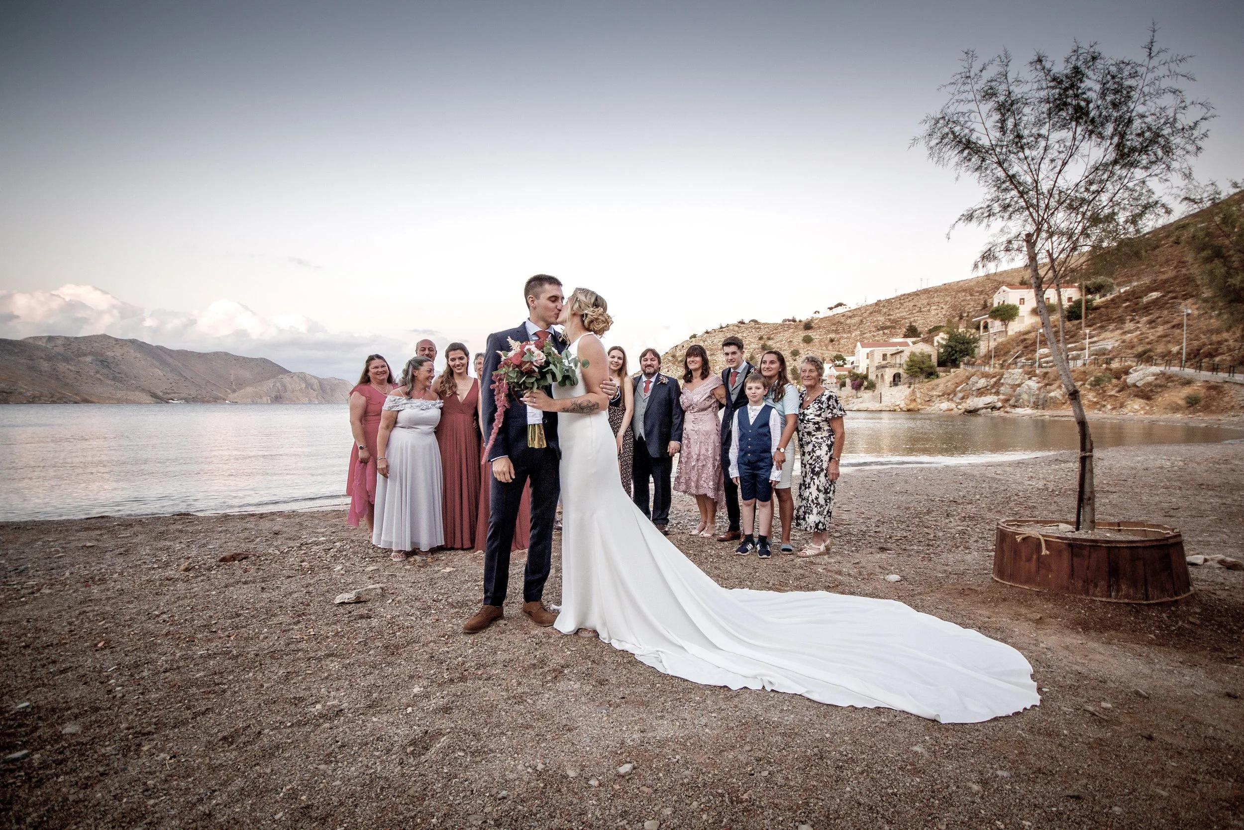 A bride and groom share a kiss during their outdoor wedding ceremony on a beach, with family and friends standing behind them, mountains, and a few houses in the background.