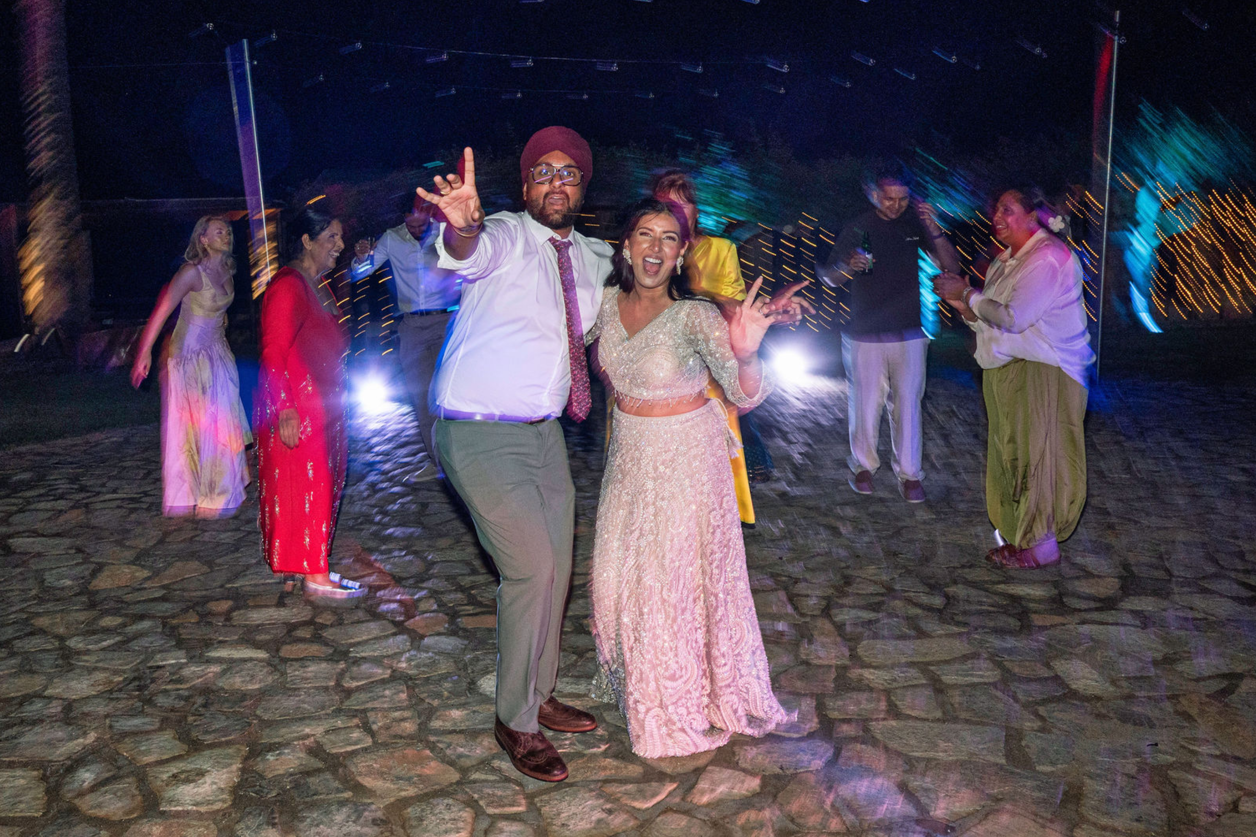 Group of people dancing and celebrating at night, dressed in festive clothes, with colorful lights and a cobblestone ground.