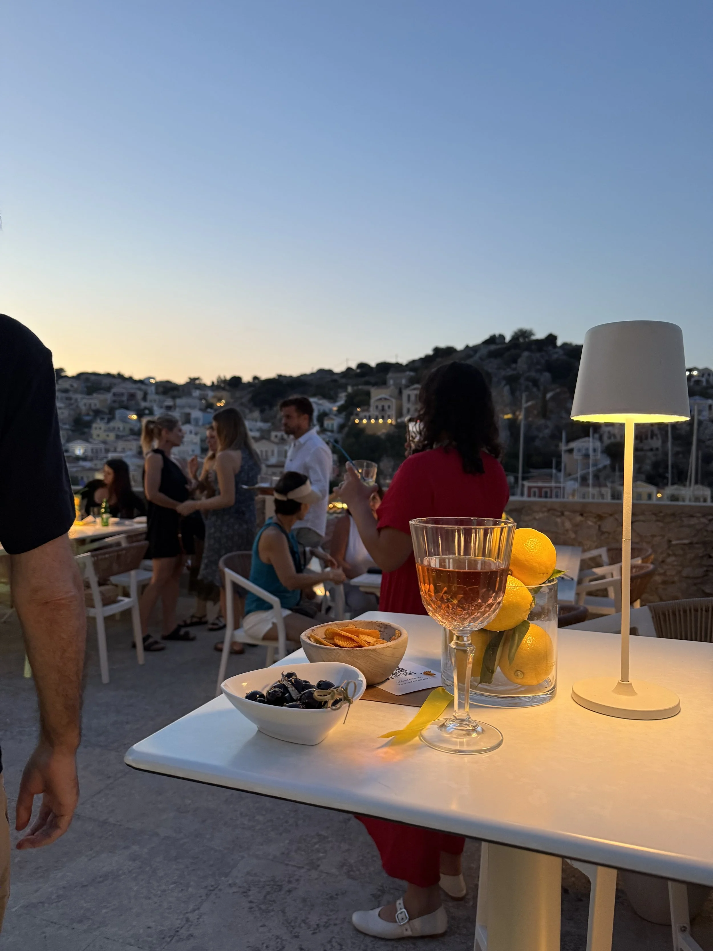 Outdoor gathering at sunset with a white table in the foreground holding a glass of rosé wine, a bowl of olives, a bowl of potato chips, a lemon, and a small toy. A woman in a red dress and white shoes is holding a phone nearby. In the background, se