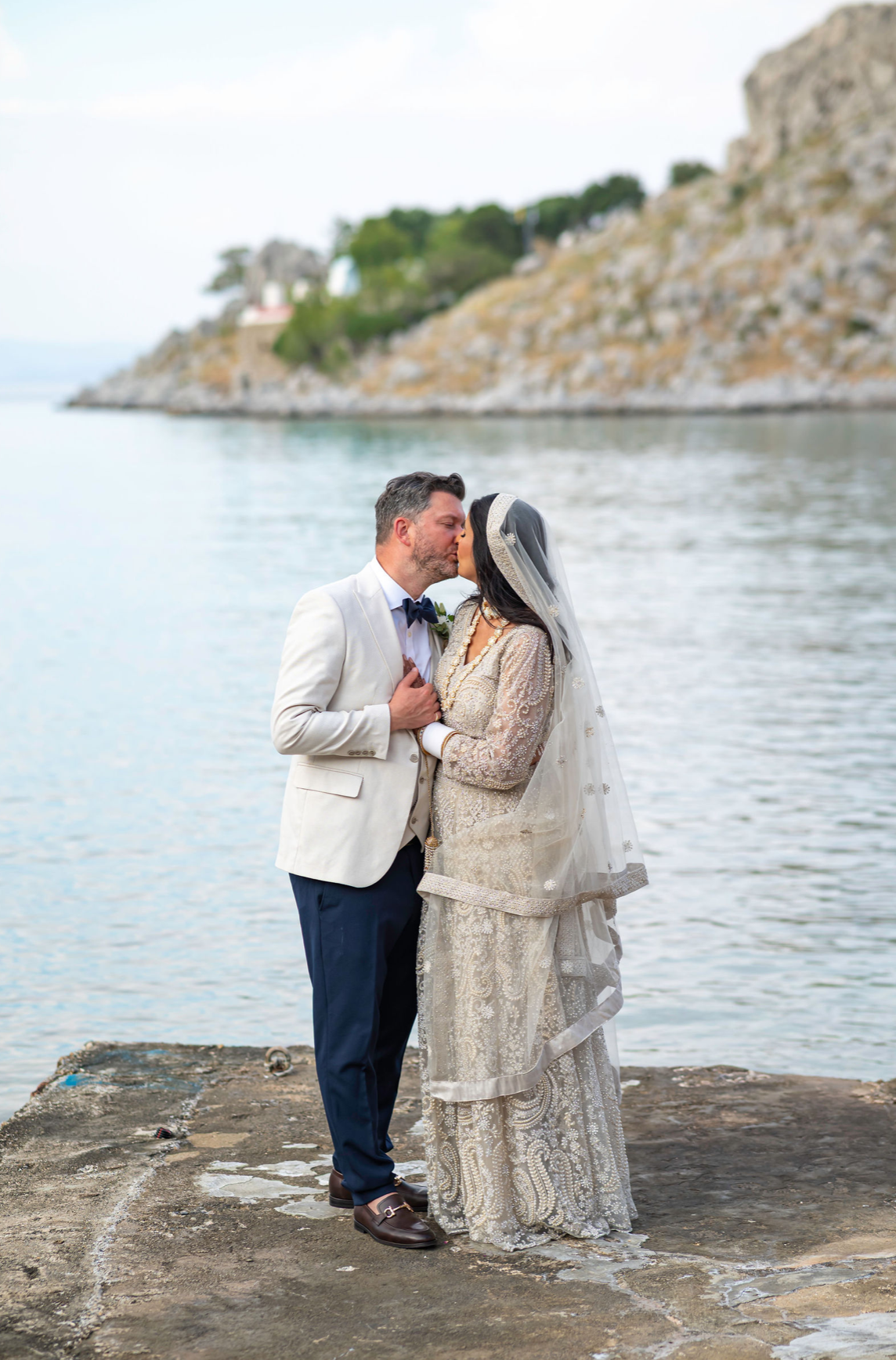 A couple dressed in wedding attire sharing a kiss by the water on a rocky pier with a hilly, rocky shoreline and a few trees in the background.
