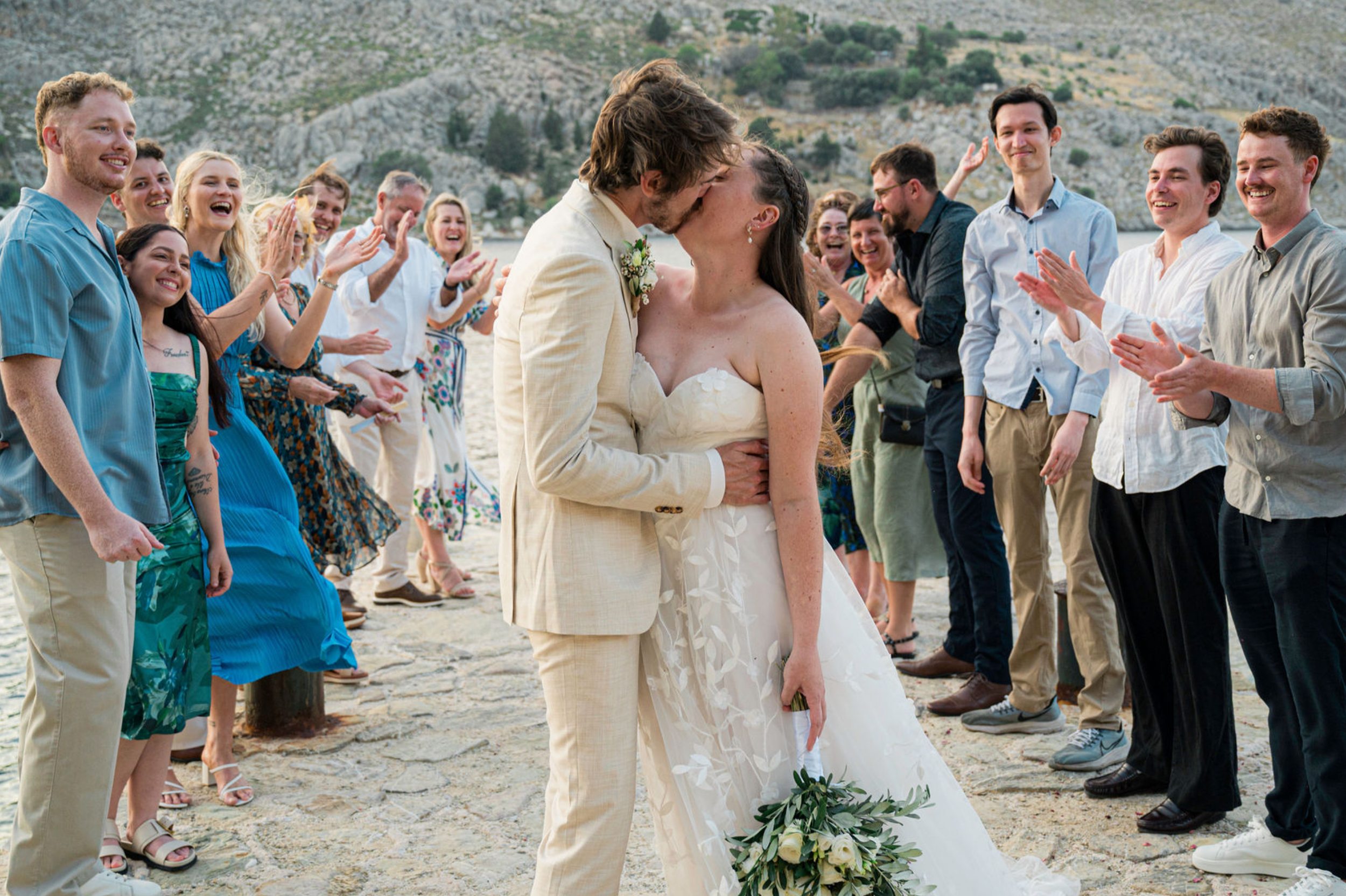 A group of people, including a bride and groom, celebrate and applaud outdoors near a body of water with mountains in the background during a wedding ceremony.