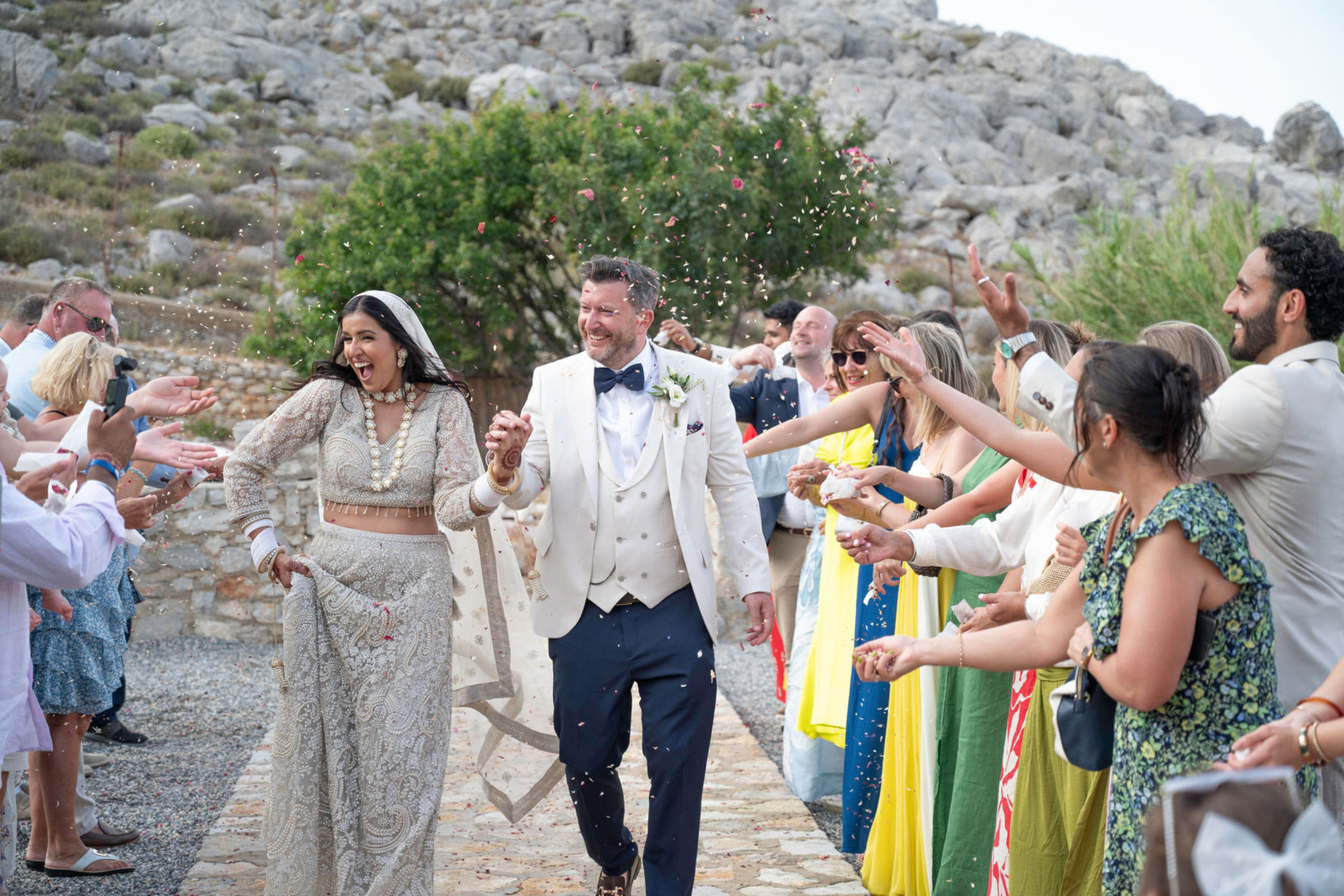 A bride and groom holding hands and smiling as they walk through a line of guests throwing confetti during a wedding ceremony outdoors near rocky terrain and green trees.