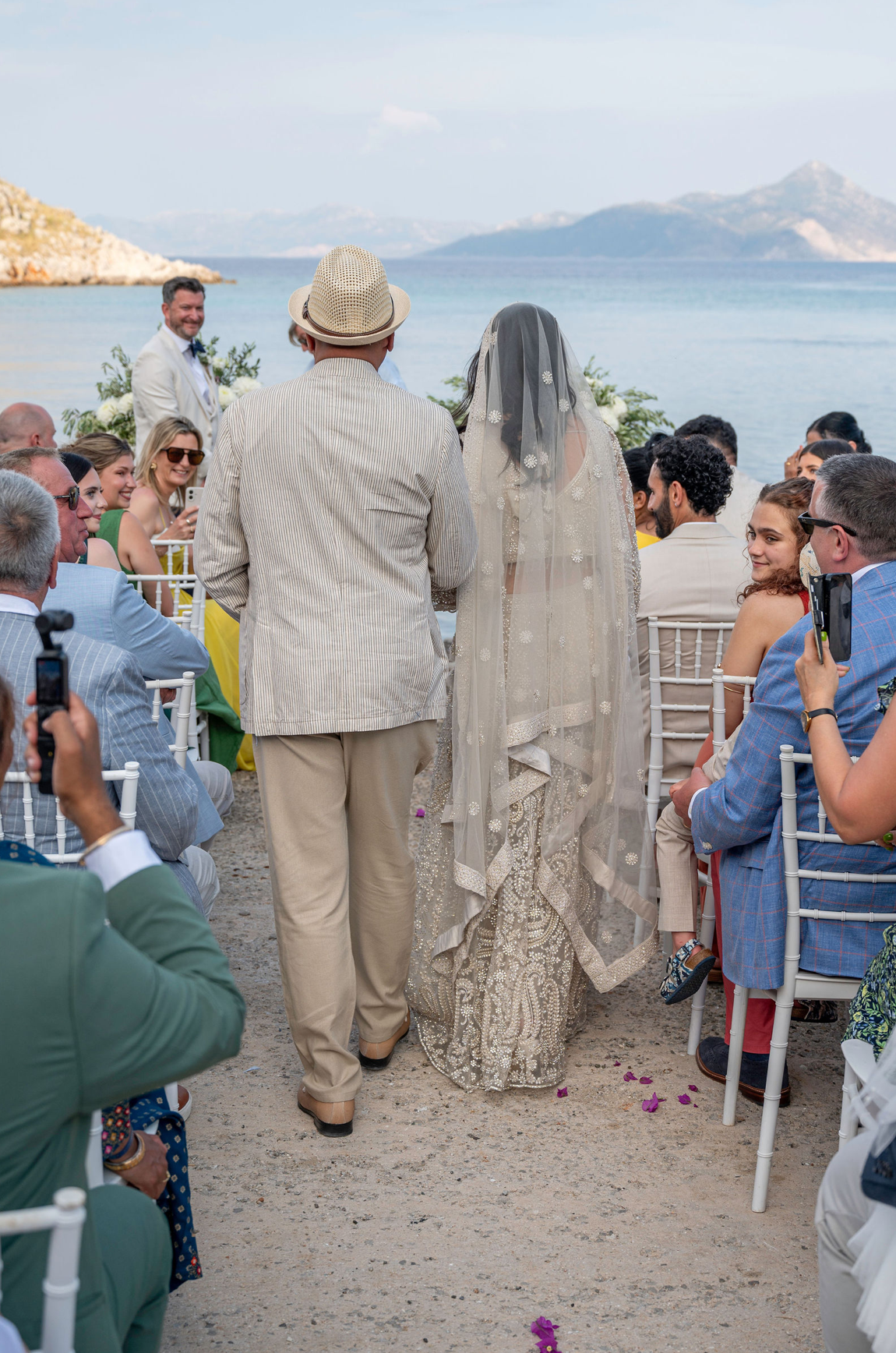 A wedding ceremony taking place outdoors by the water with a bride in a detailed lace gown and veil, walking with a man in a striped suit and hat, while guests are seated and watching.