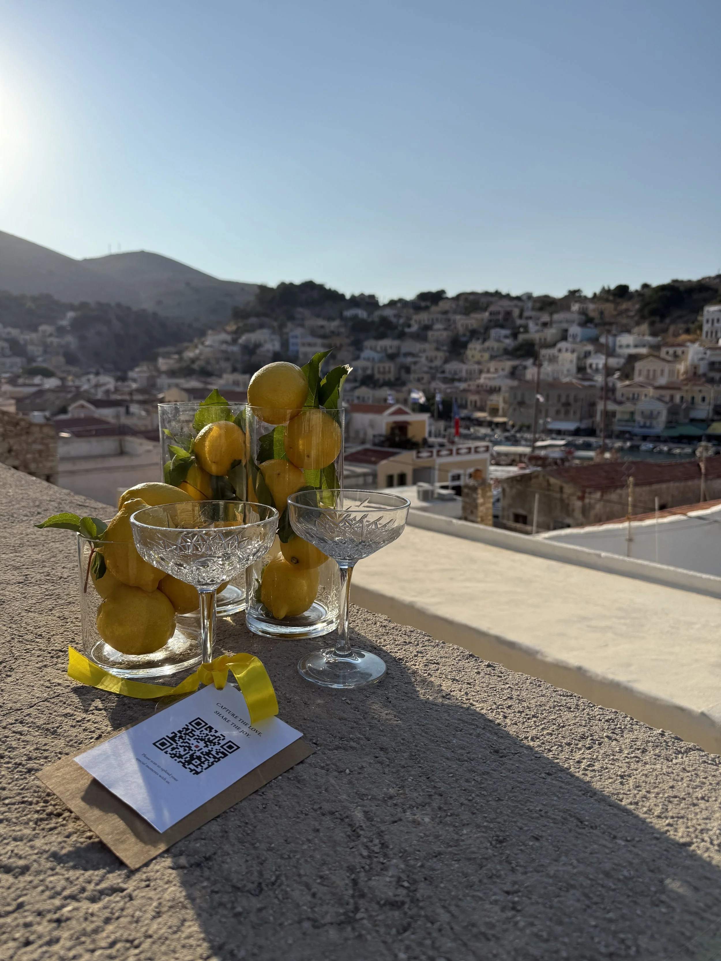 Glass vases filled with yellow lemons and green leaves, set on a ledge overlooking a coastal town with white buildings and rolling hills, in bright sunlight.