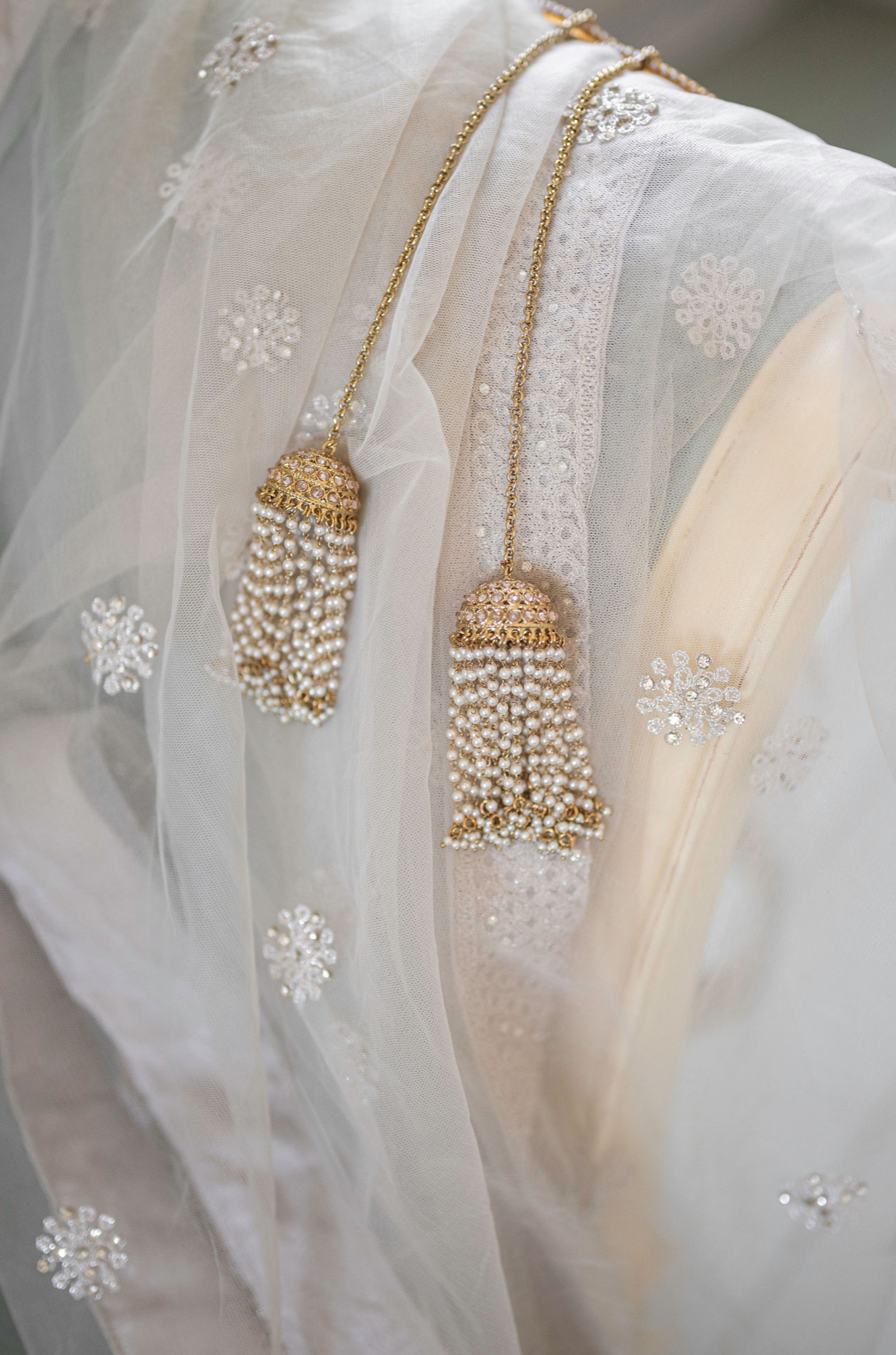 Close-up of a pair of traditional Indian earrings with gold chains and pearl tassels hanging over sheer fabric with embroidered floral patterns.